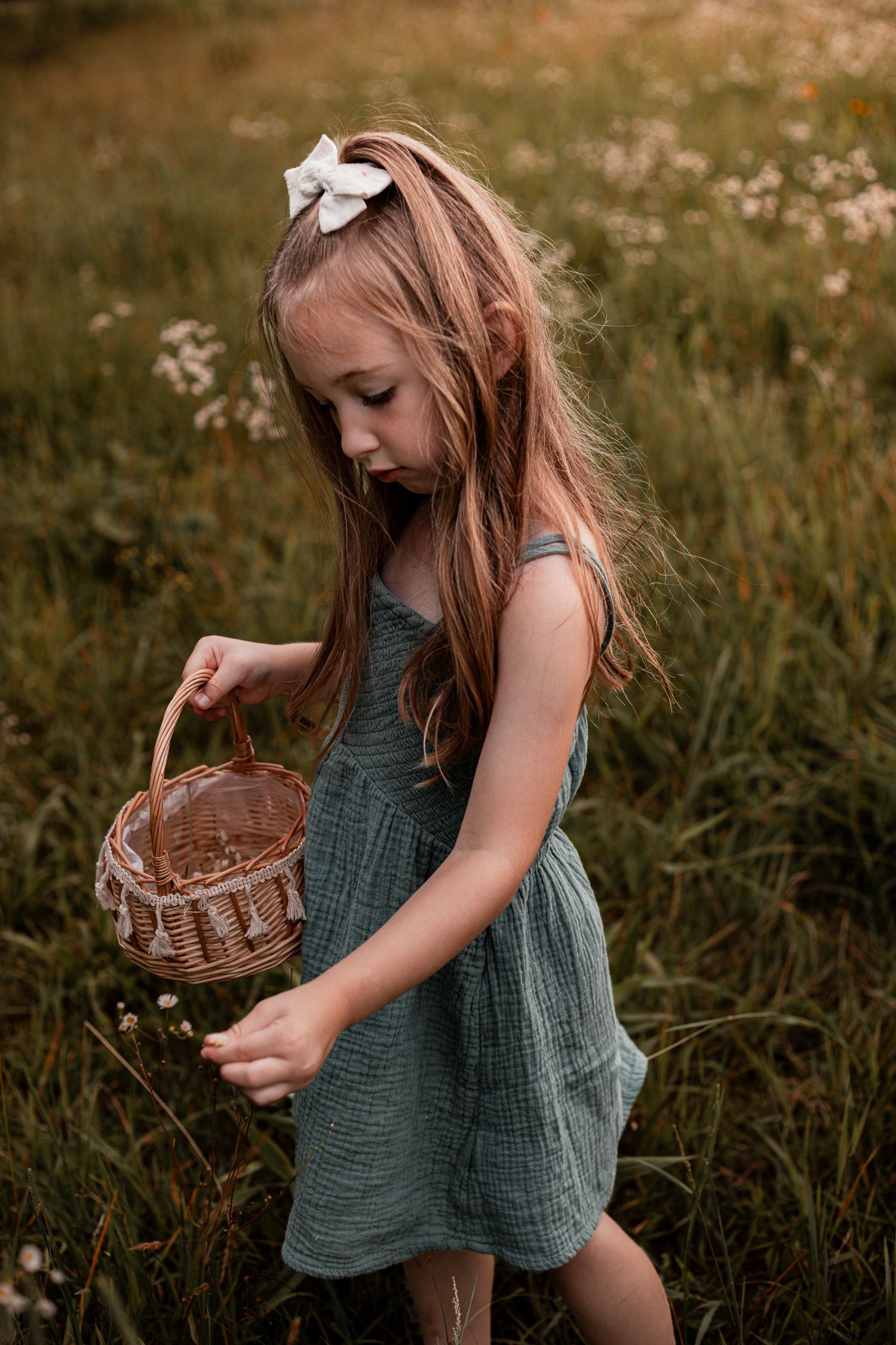 A young girl in a green dress carries a wicker basket while exploring a wildflower meadow at golden hour.