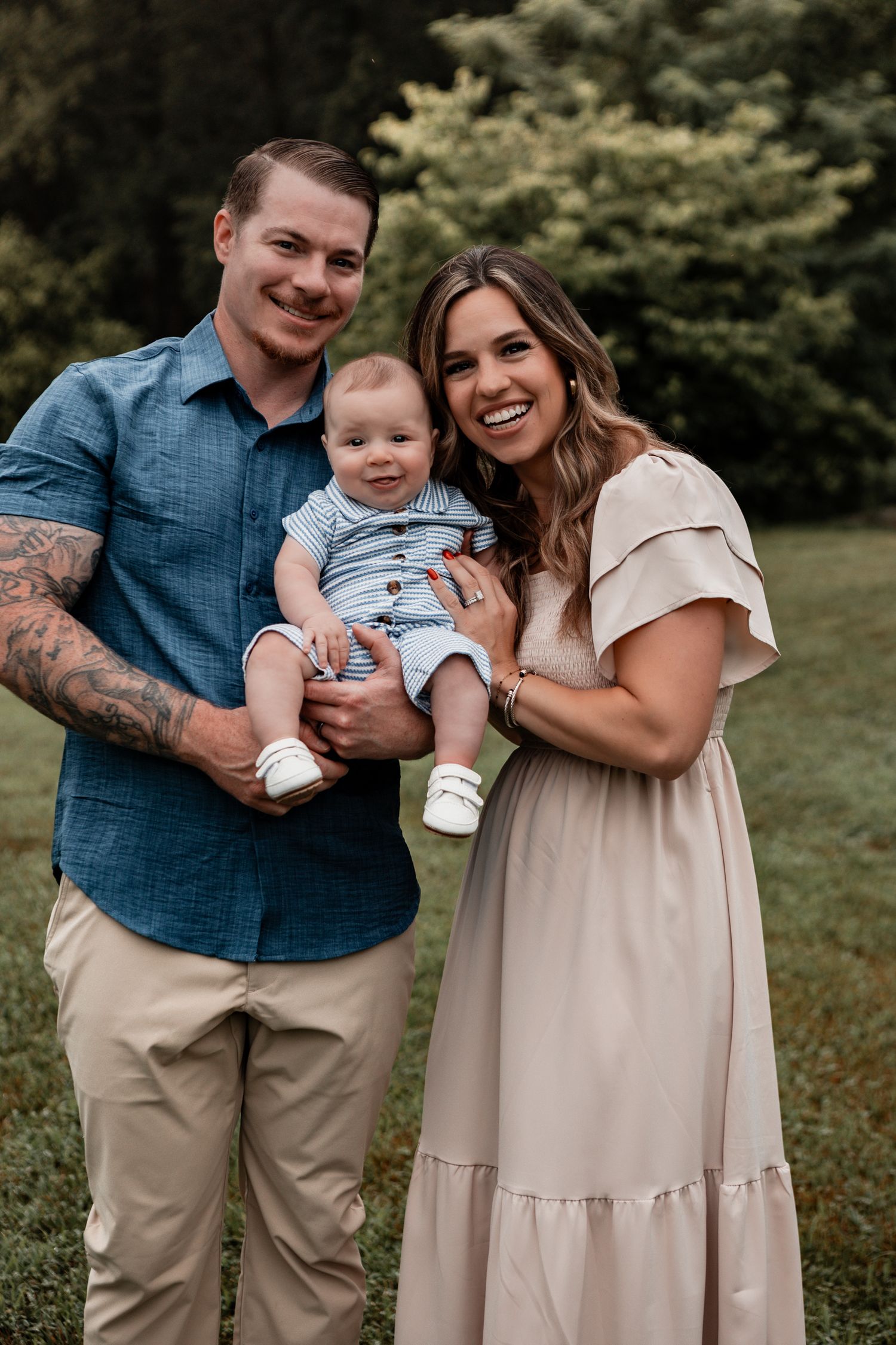 A family laughs together in an outdoor setting wearing coordinated beige and blue outfits against a lush green backdrop.