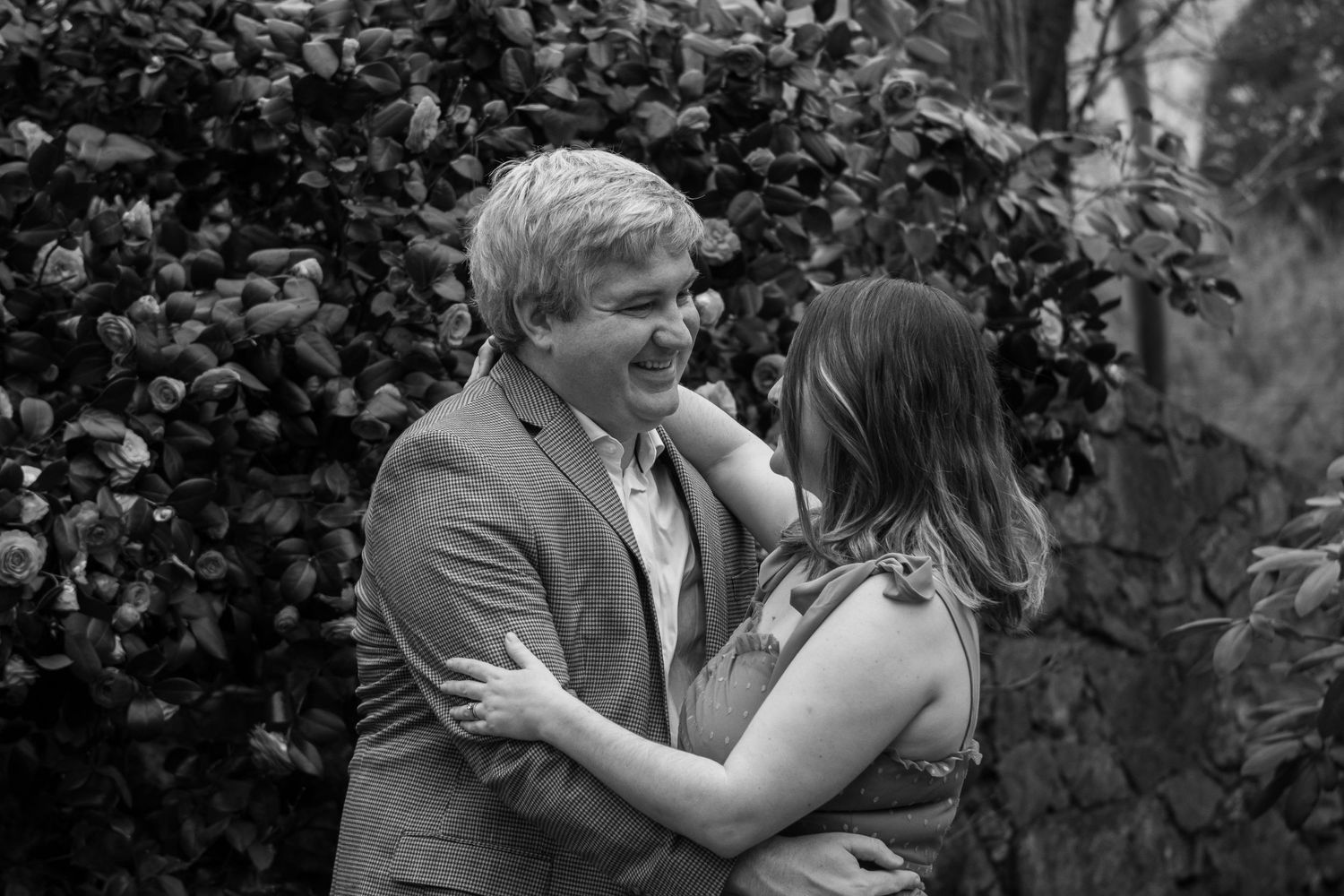 Black and white romantic moment of a couple embracing and laughing together against a garden backdrop.