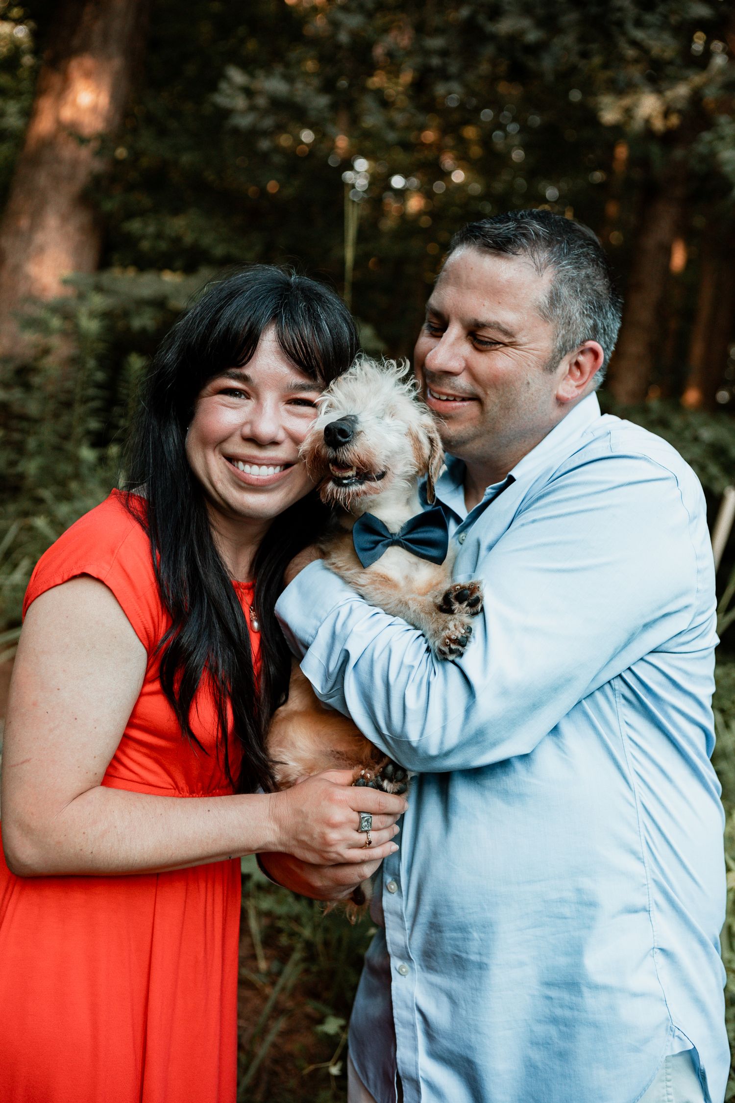 A couple in red and light blue clothing share joyful moments with their small pet dog during a forest photoshoot.