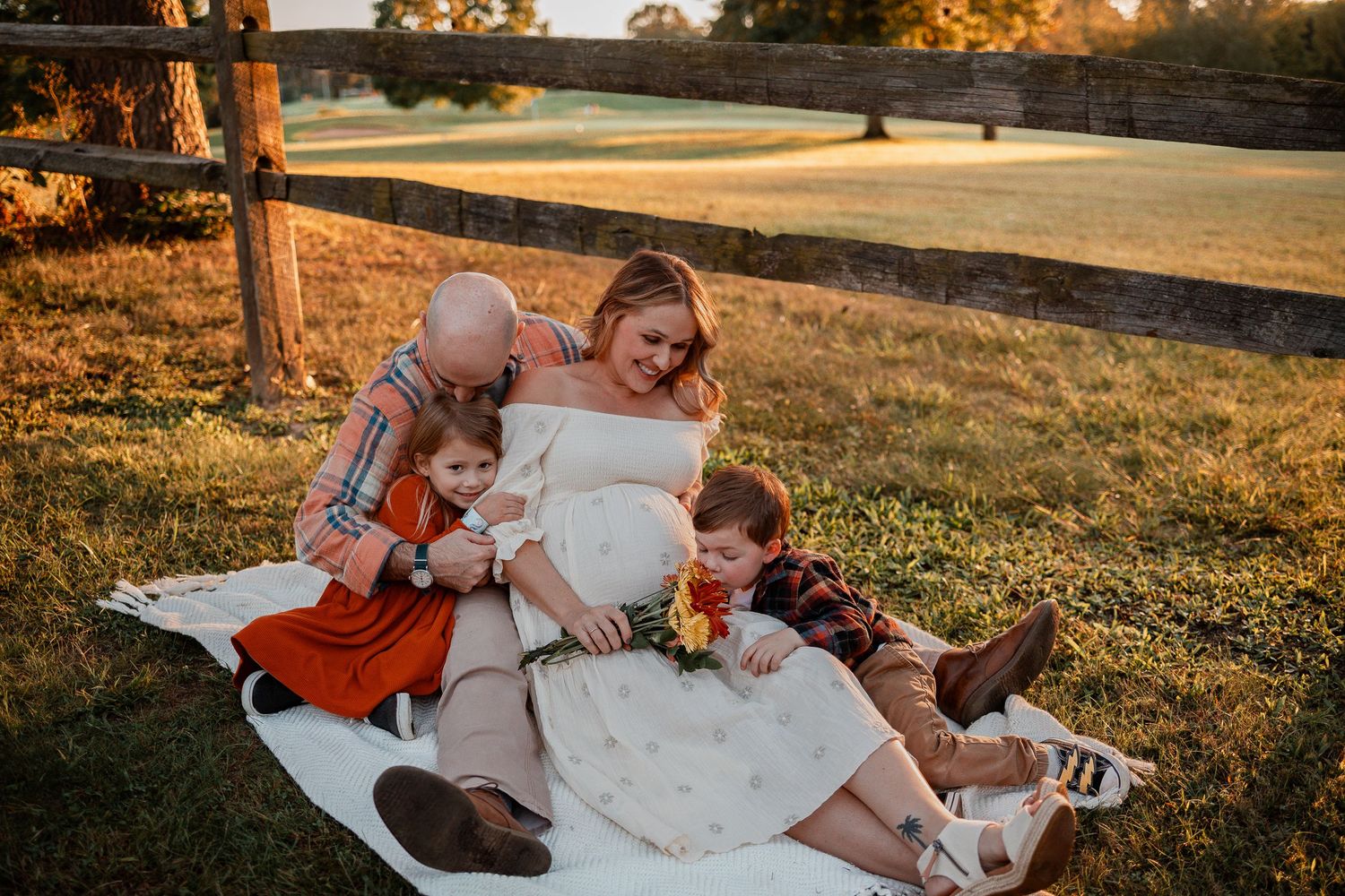 A pregnant woman in a white dress sits on a blanket outdoors at golden hour with two young boys leaning against her.