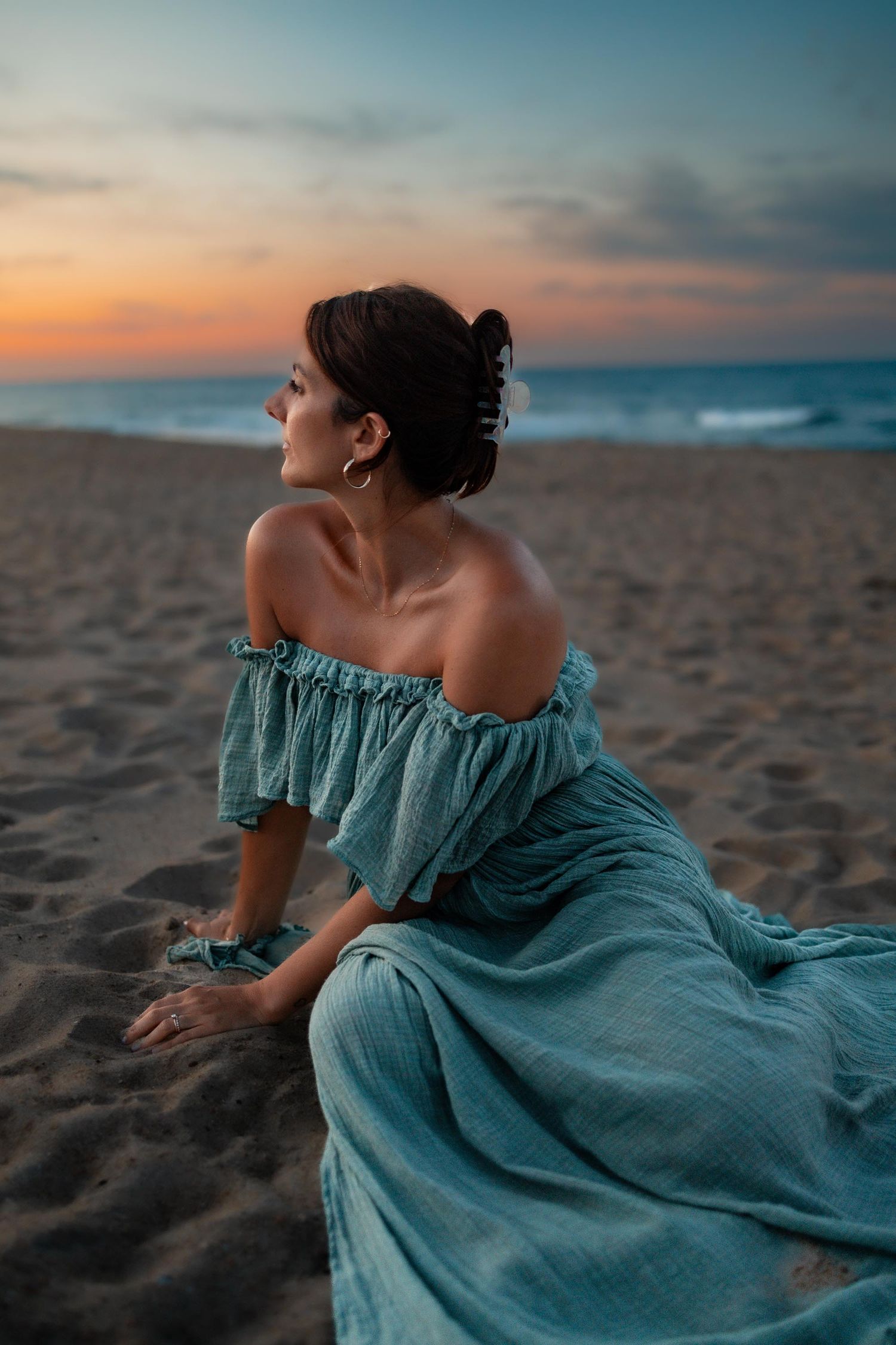 Woman in teal off-shoulder dress sitting on sandy beach at sunset, gazing toward colorful ocean horizon.