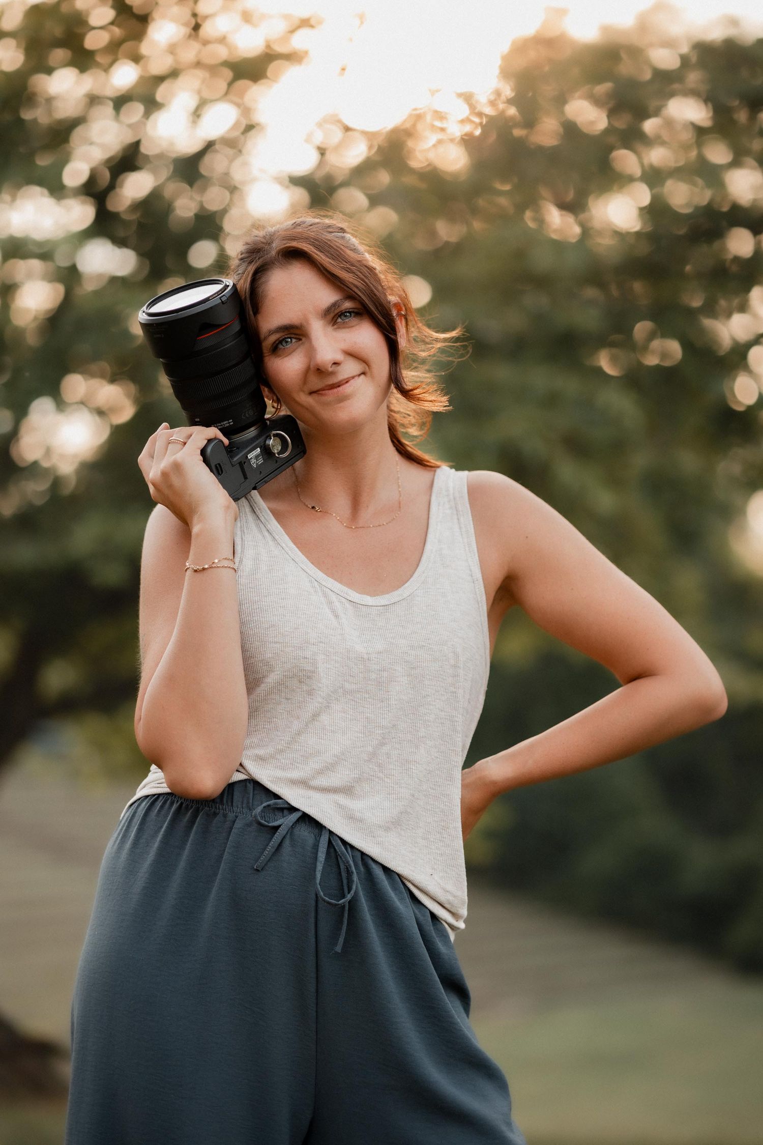 Woman photographer holding DSLR camera outdoors at golden hour, wearing white tank top and teal pants.