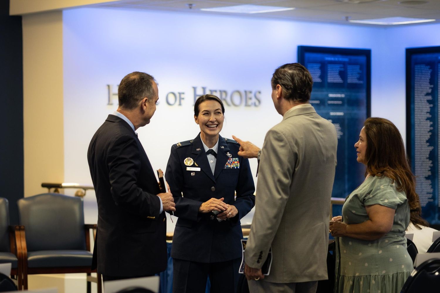 Military officer in uniform conversing with civilians in front of a Hall of Heroes display at a formal event.