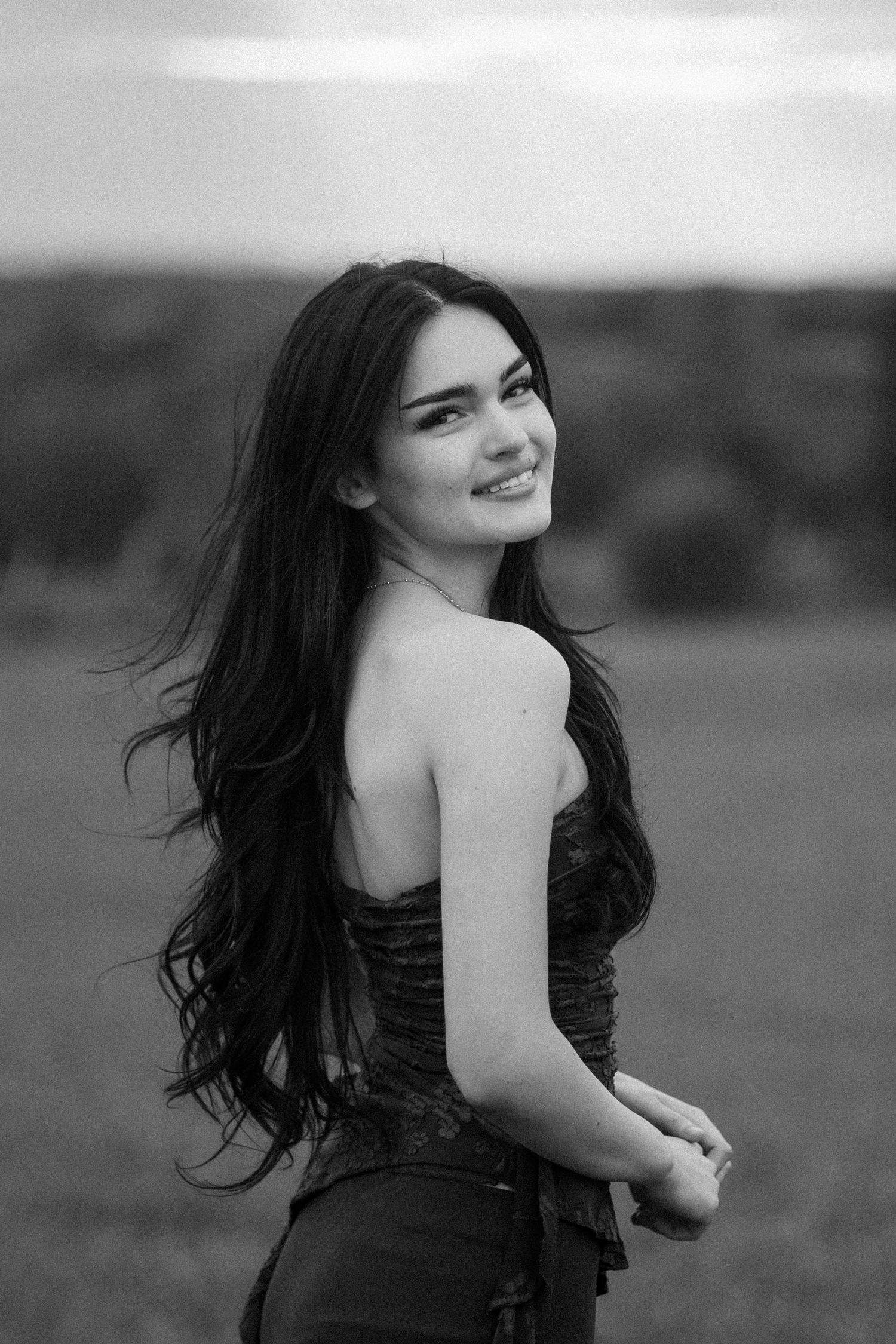Black and white portrait of a smiling young woman with long dark hair in a strapless top outdoors.