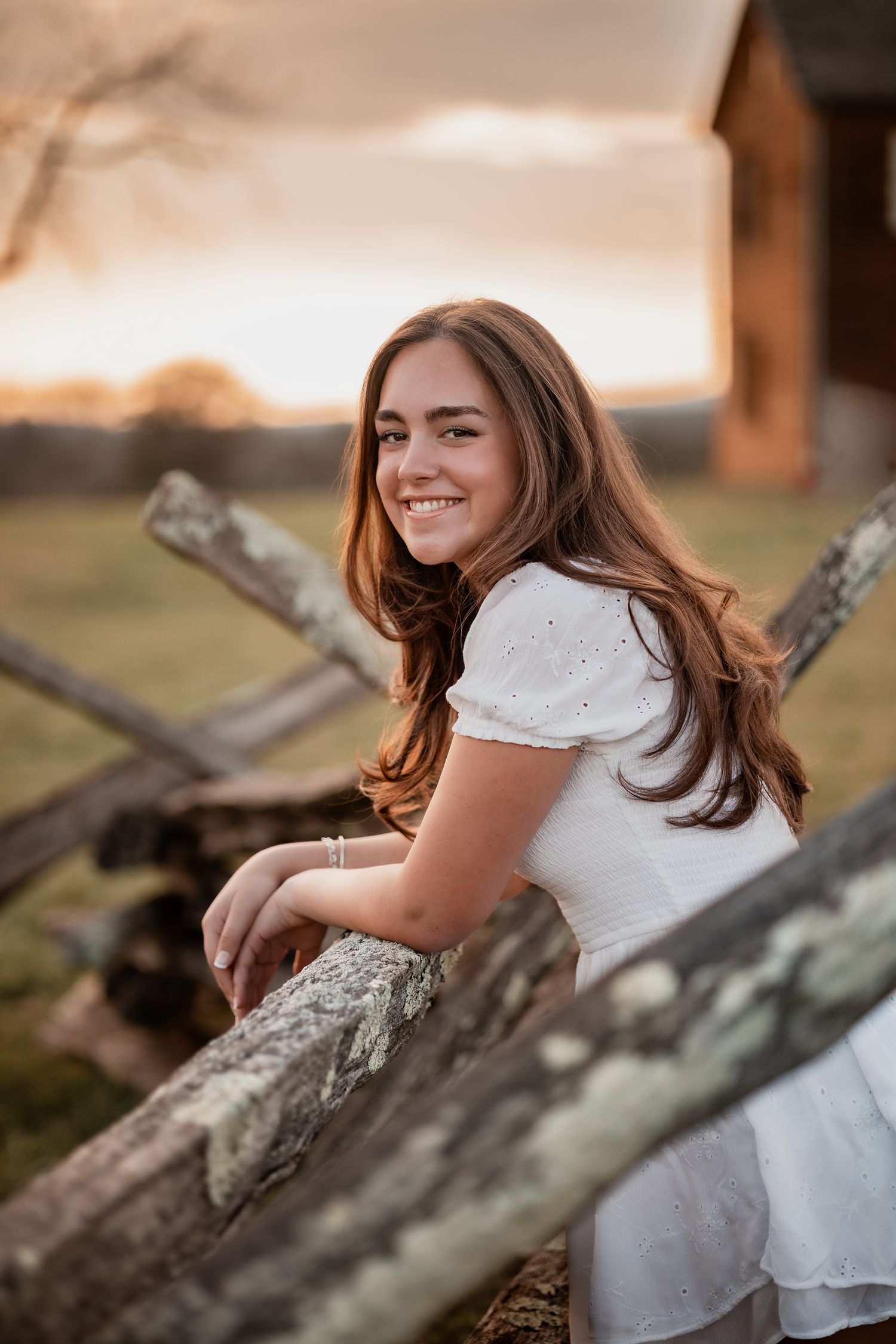 Smiling teenage girl in white dress leaning on rustic wooden fence at golden sunset near a barn.