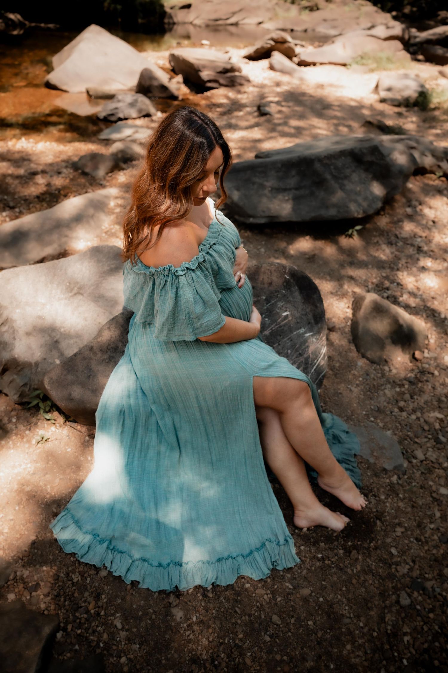 Pregnant woman in teal off-shoulder dress sitting barefoot on rocks in a serene outdoor setting.