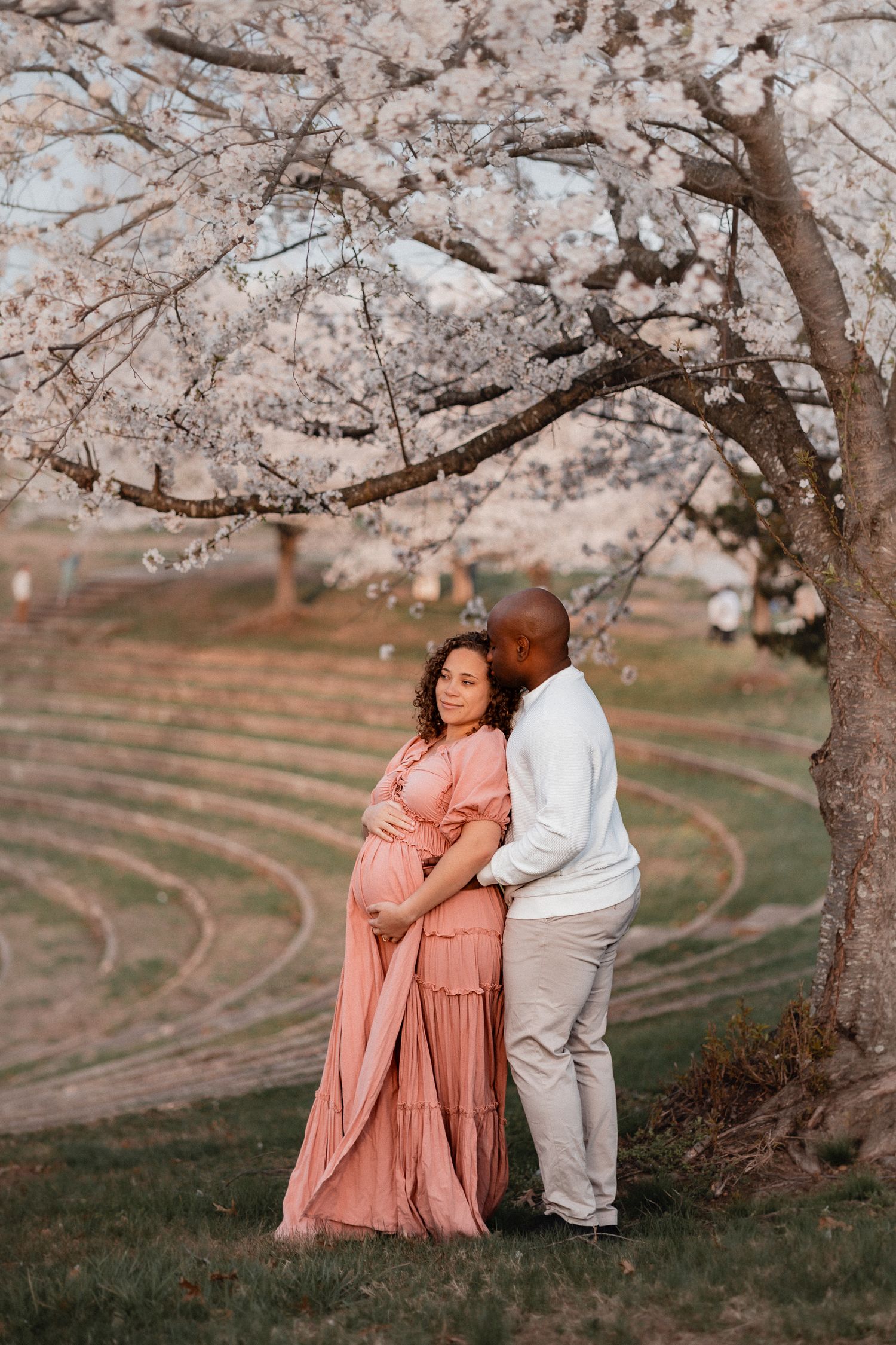 Pregnant couple embracing under blooming cherry blossom tree, woman in pink maxi dress, man in white sweater, spring maternity photo.