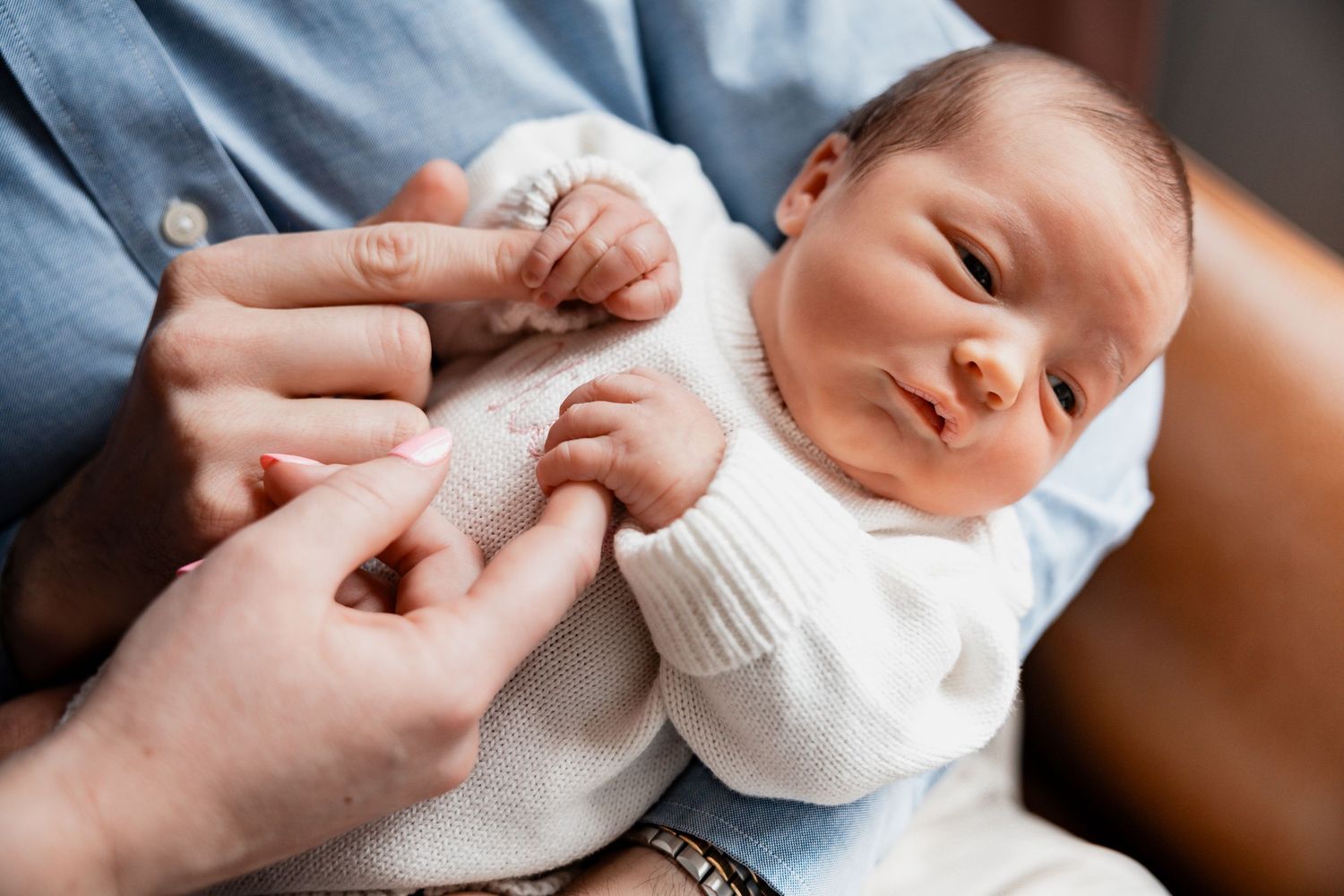 Newborn baby in white knit outfit being cradled in adult arms, with tiny fingers visible.