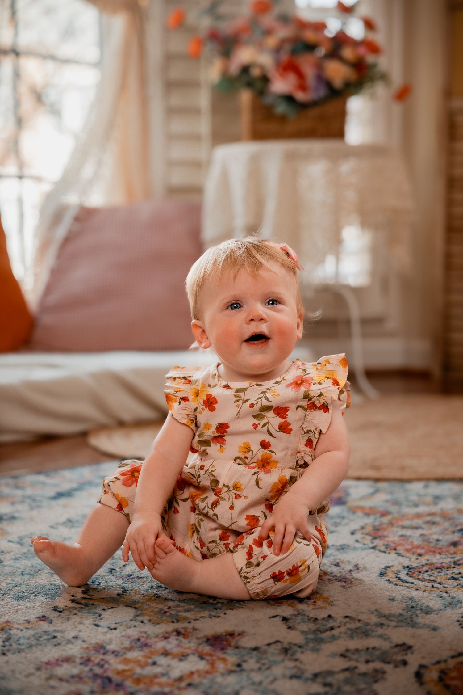 Smiling baby girl in floral romper sitting on a rug in a cozy, warmly decorated living room.