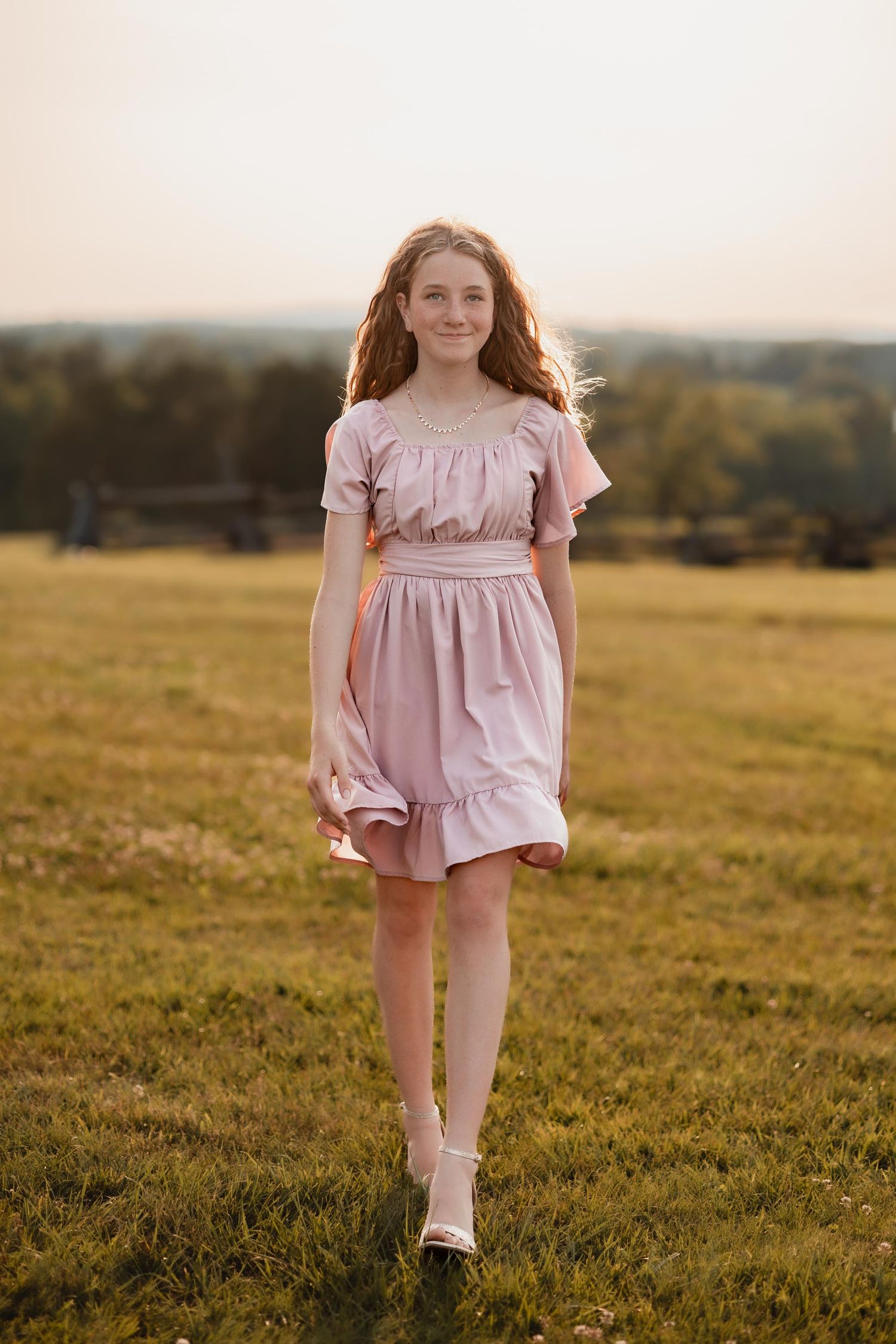 Young girl in pink ruffle dress standing in a sunlit green field at golden hour, smiling with curly hair flowing.