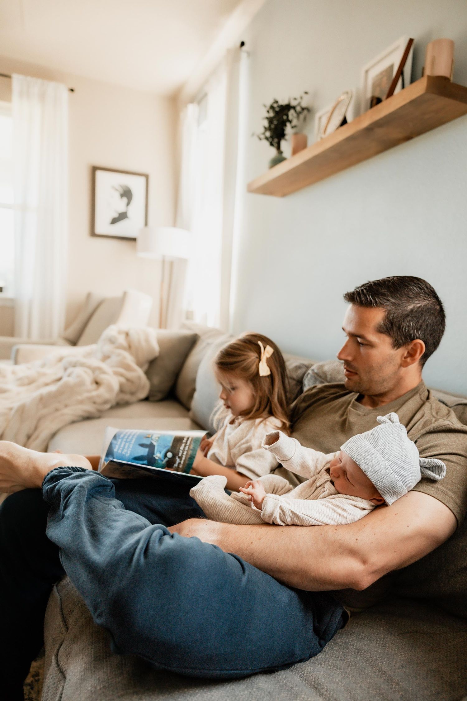 Father sits on floor holding newborn baby while toddler daughter reads a book beside him in cozy living room.