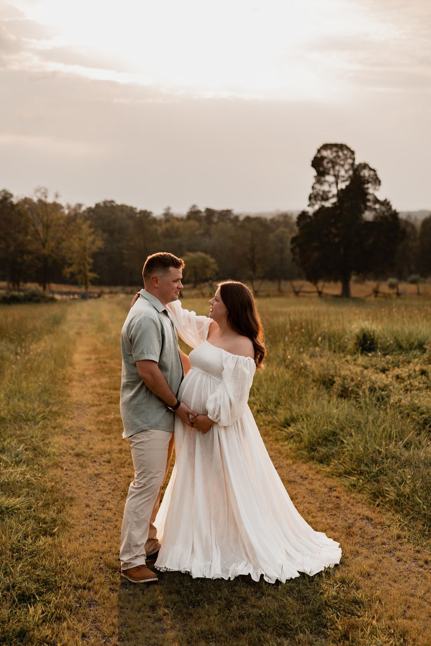 Couple sharing a tender moment on a rustic path in a golden field at sunset, woman in flowing white maternity dress.