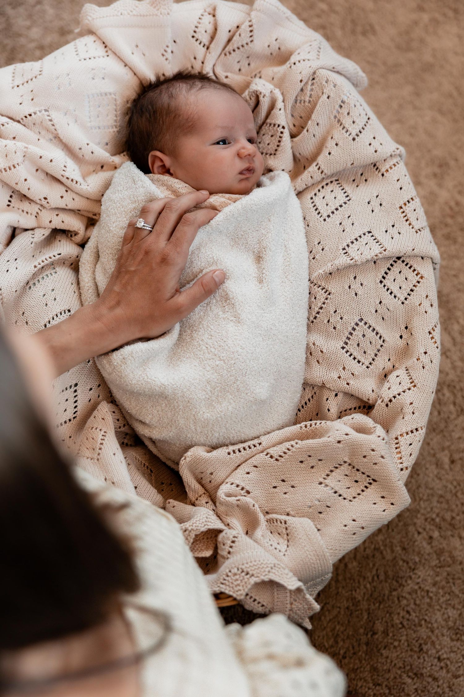 Newborn baby swaddled in white knit blanket resting in wicker bassinet with mothers hand nearby.