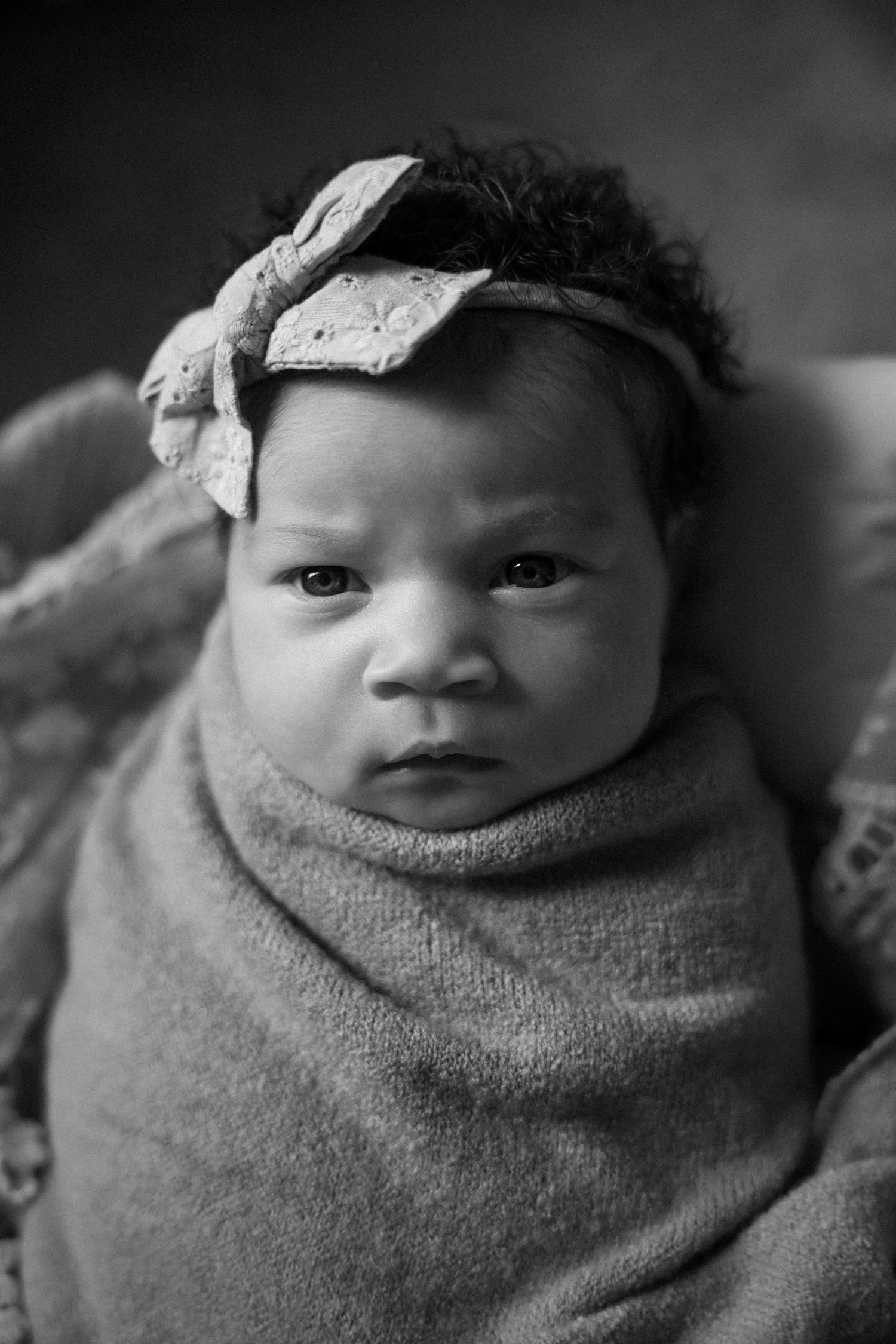 Black and white photo of a swaddled newborn baby girl wearing a bow headband, looking alert and curious.