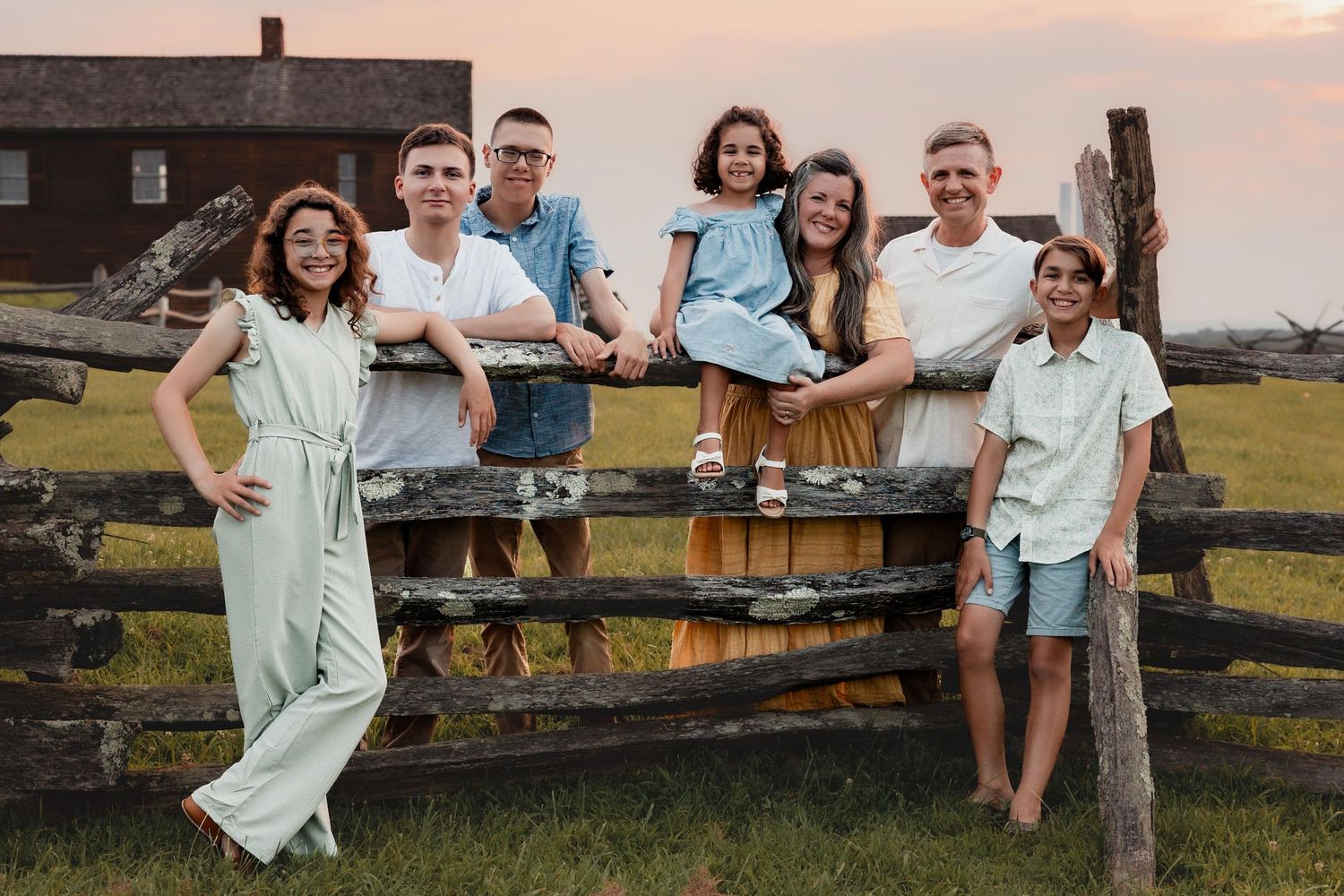 Happy family of seven posing together by a rustic wooden fence in a green meadow at sunset.