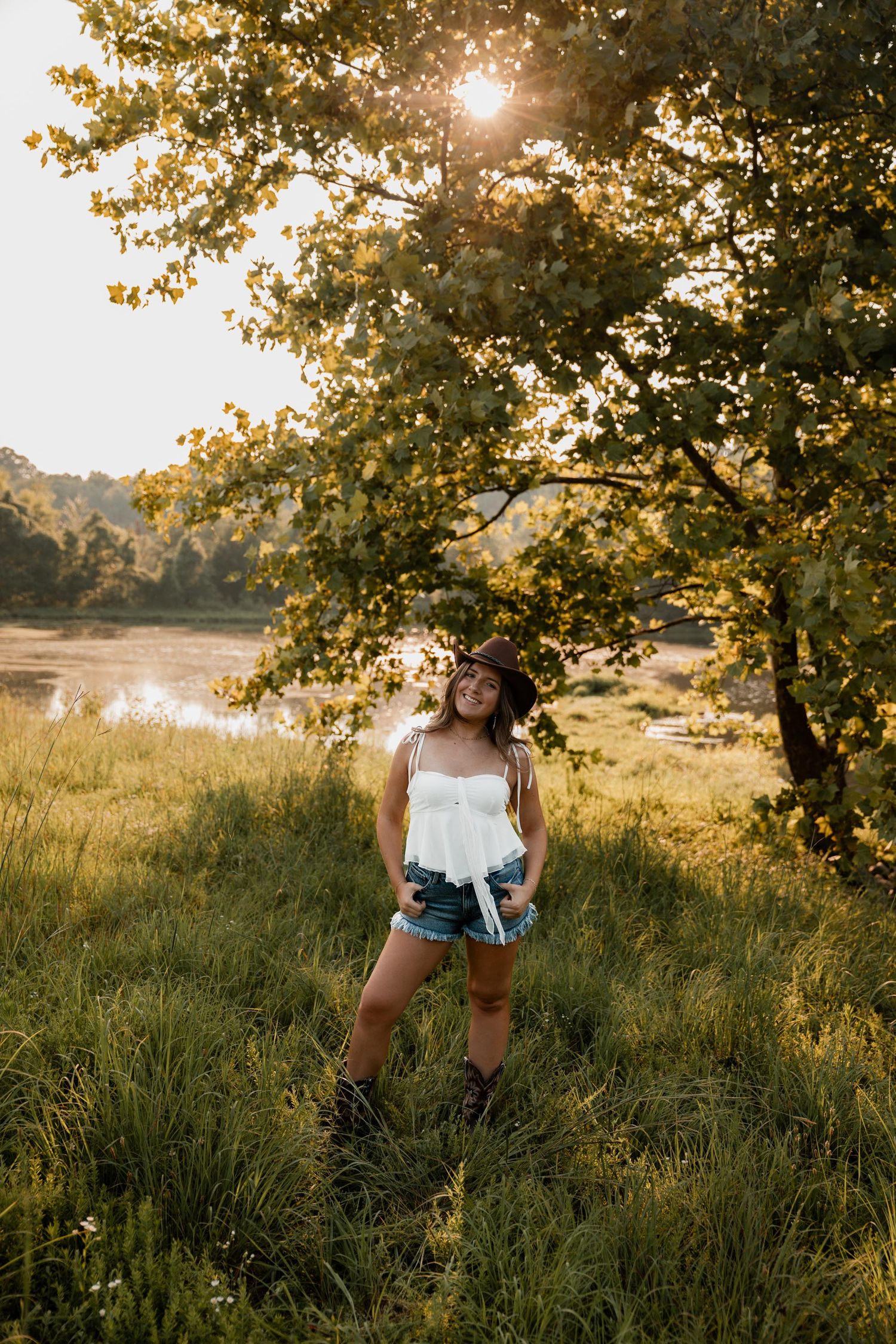 Young woman in white top, denim shorts, and cowboy hat standing in golden sunlit field near a lake under a large tree.