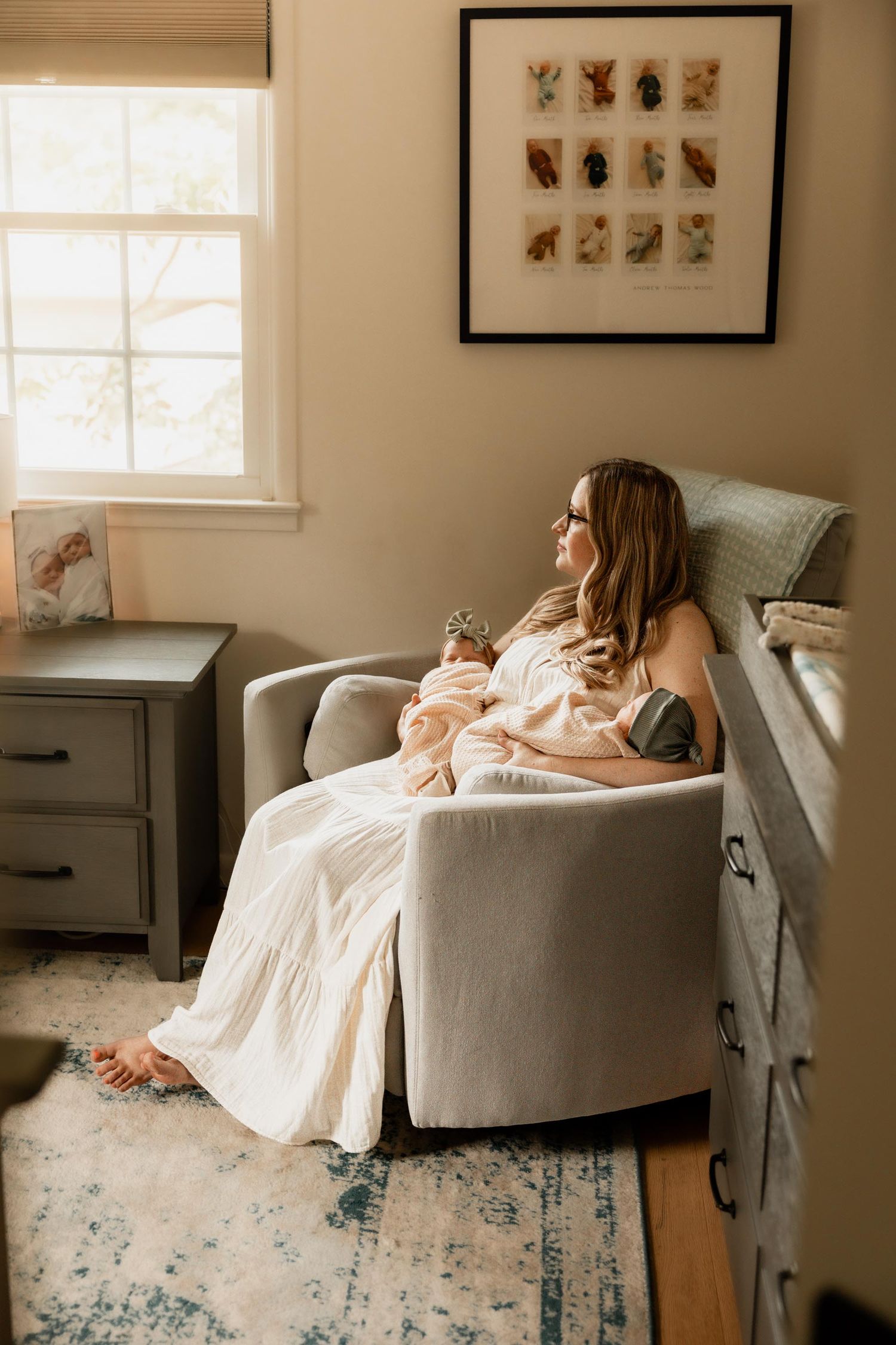 A woman in a white dress sits in a nursery glider chair, cradling her newborn baby near a sunlit window.