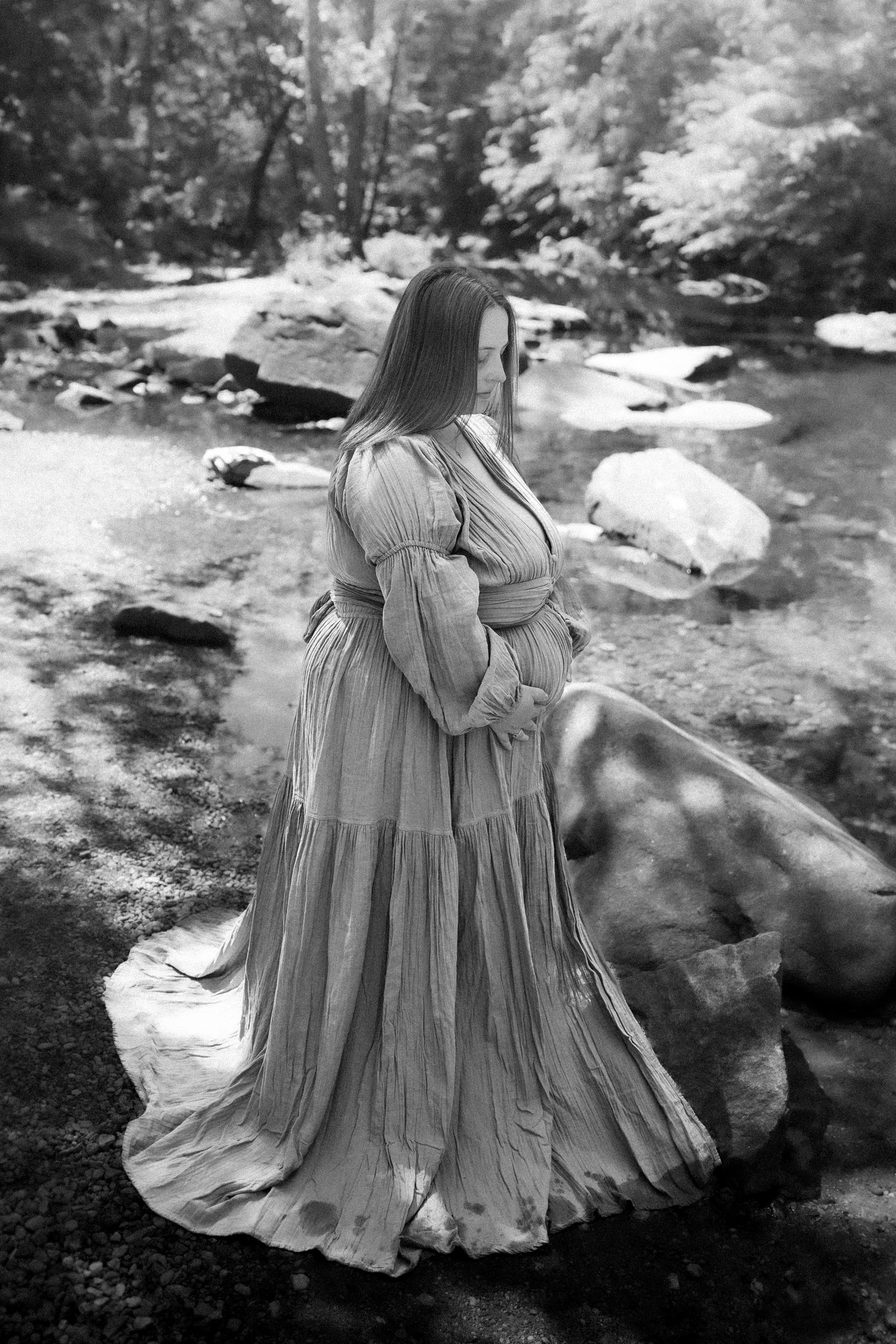 Black and white photo of a pregnant woman in a flowing dress standing by a rocky creek surrounded by trees.