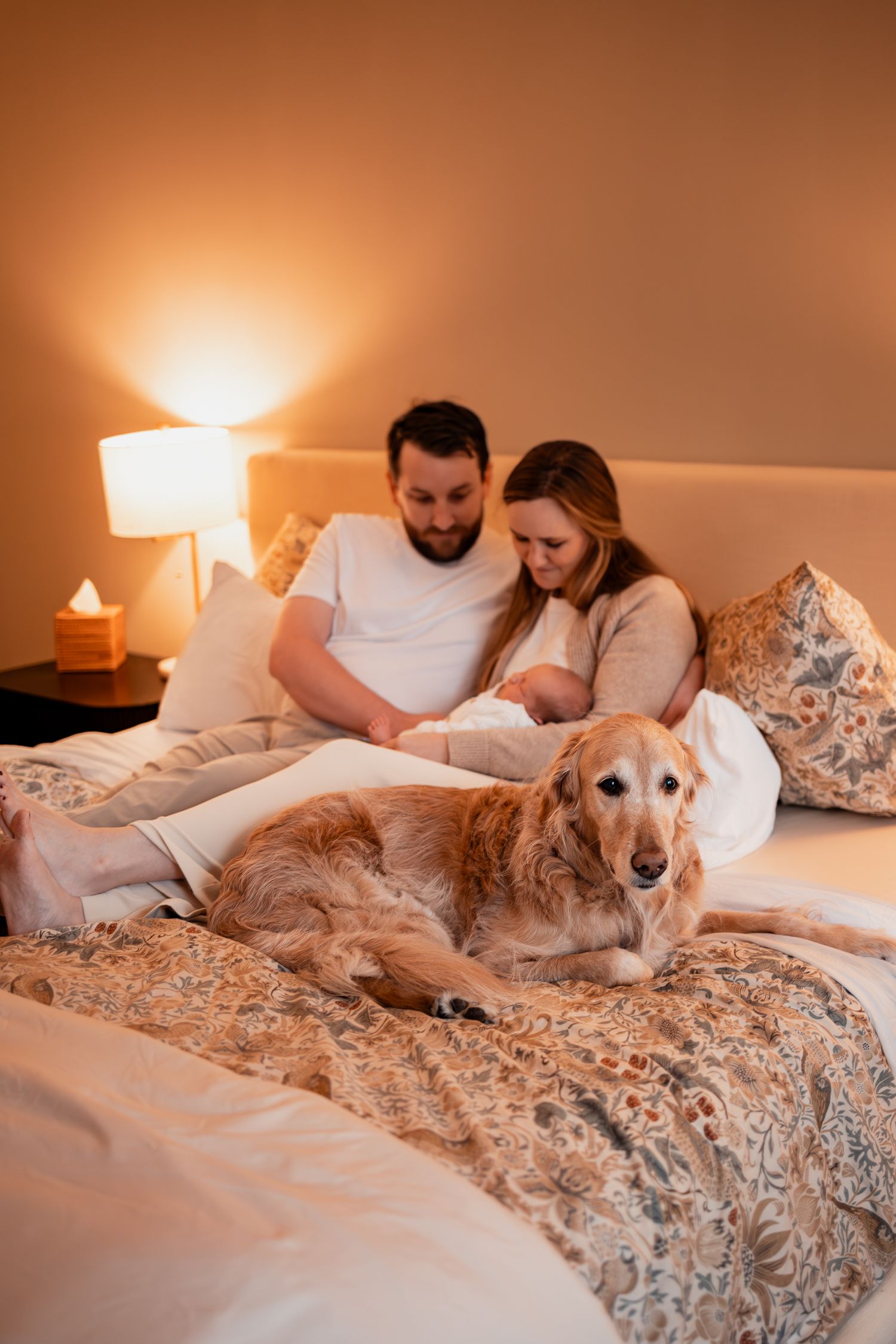A golden retriever lounges on a floral bedspread with its family in a warmly lit bedroom.
