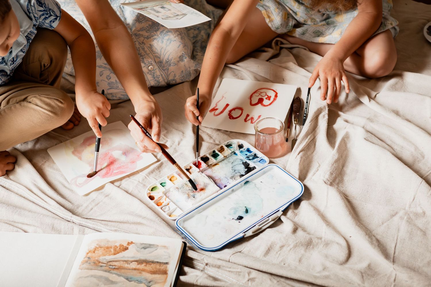 Hands holding paintbrushes over a watercolor palette and paper while painting on white bedsheets.