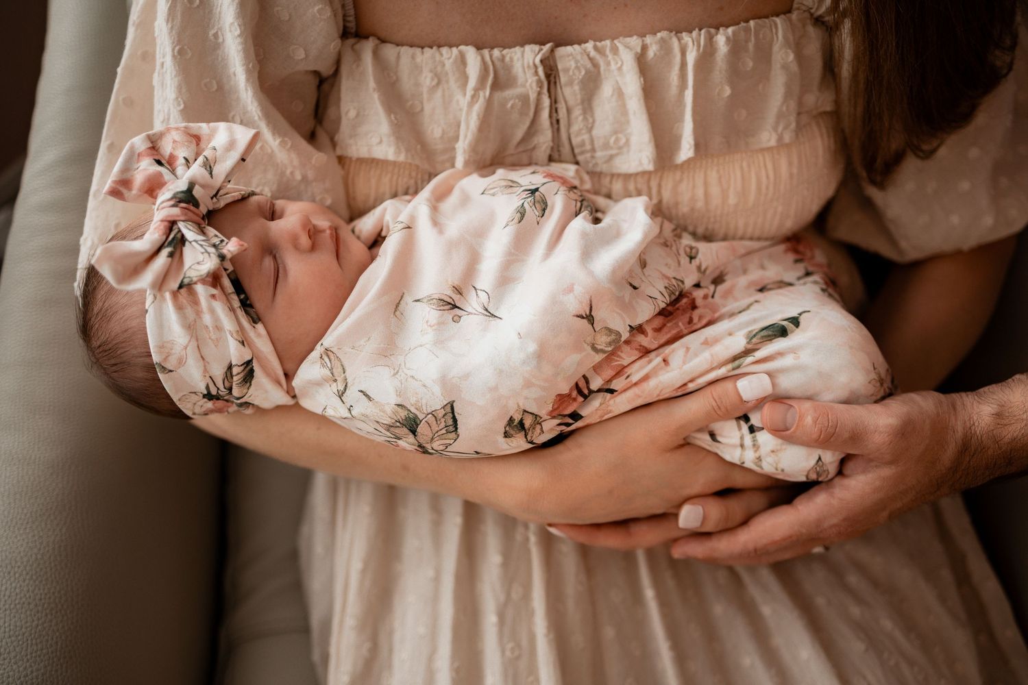 Hands cradle a sleeping newborn baby wrapped in soft white fabric during a tender portrait session.