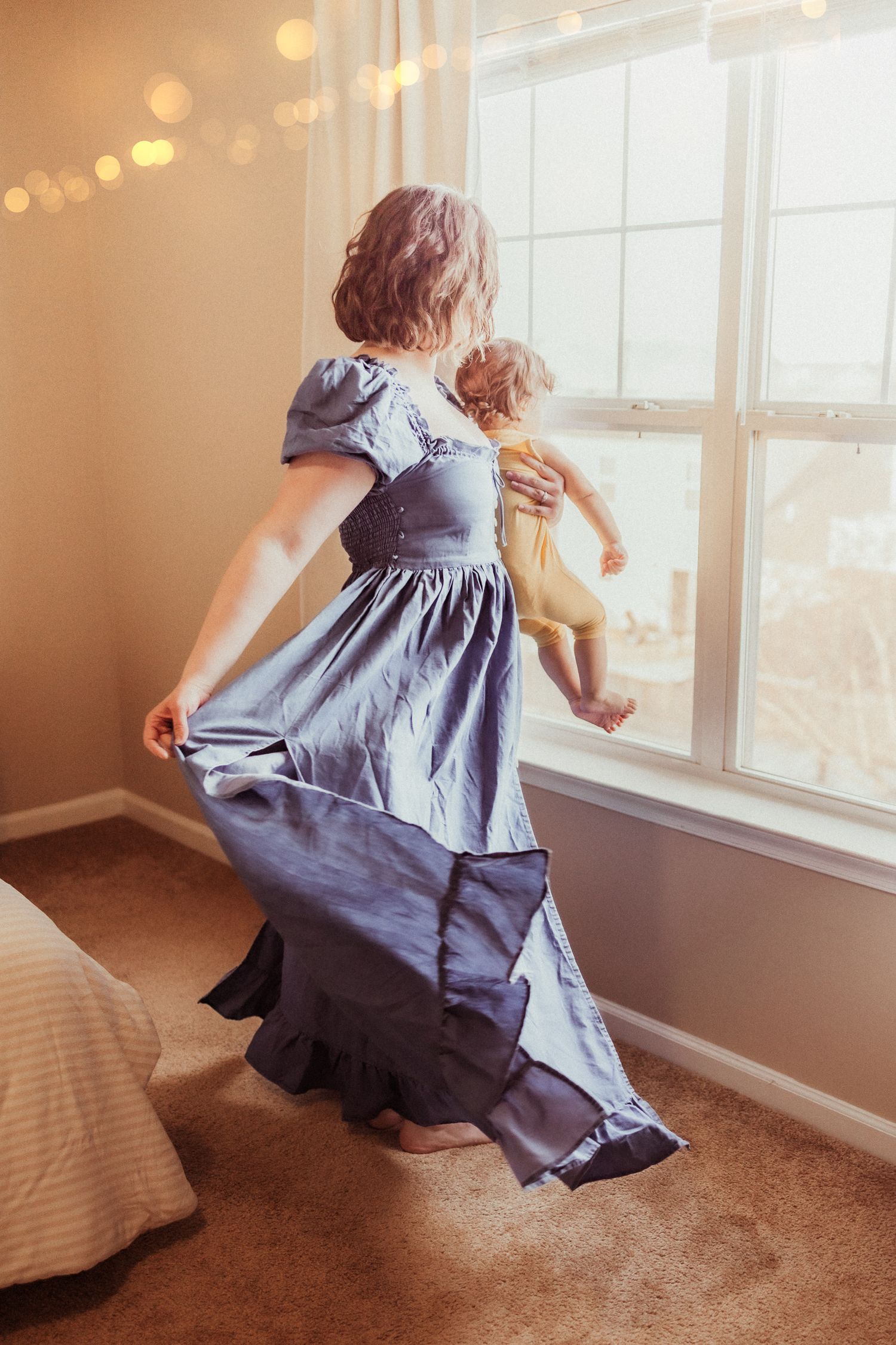 A person in a flowing blue dress twirls with a baby by a window with string lights in a warm-lit room.