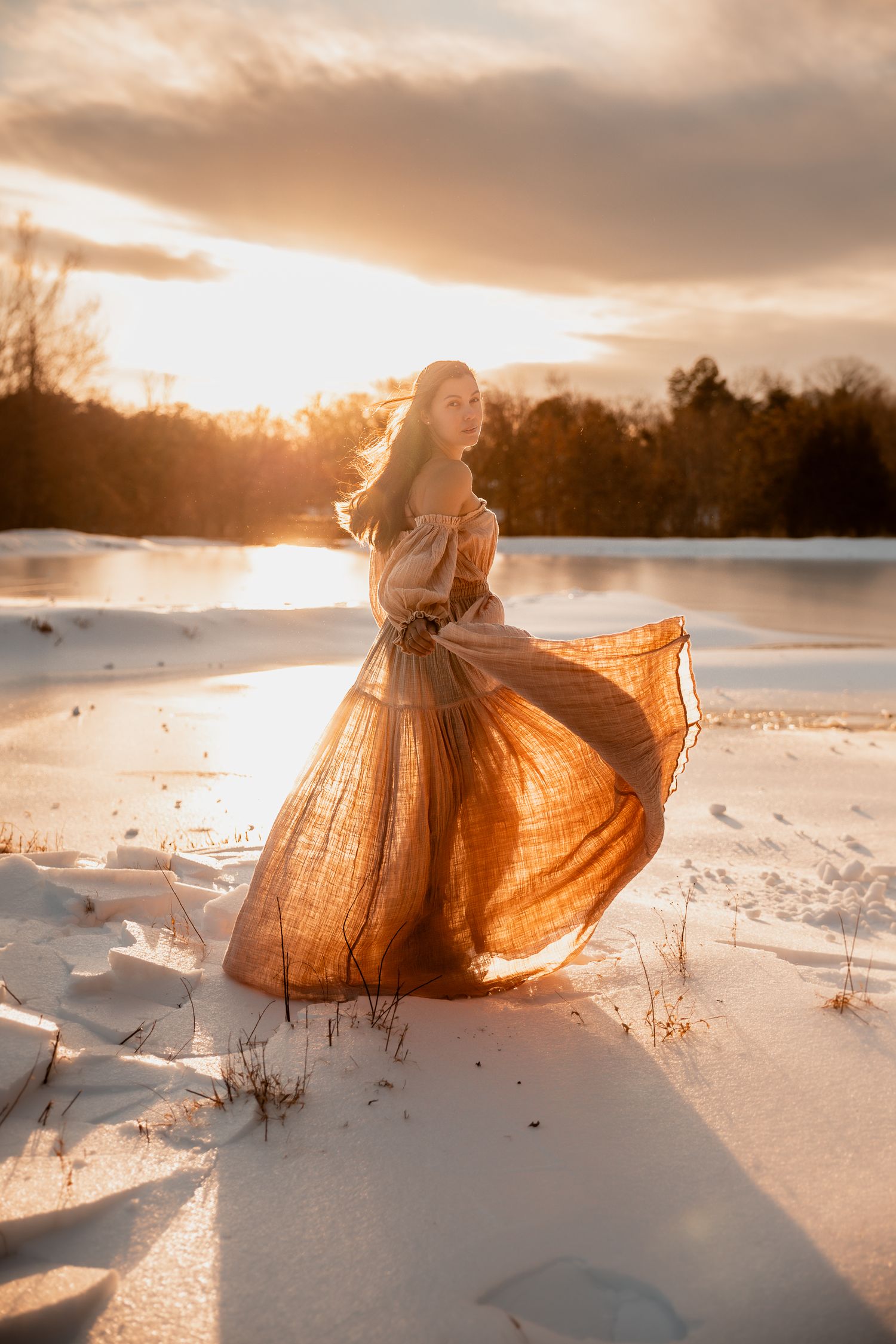 A flowing orange dress billows in the wind against a snowy landscape at sunset creating an ethereal silhouette.
