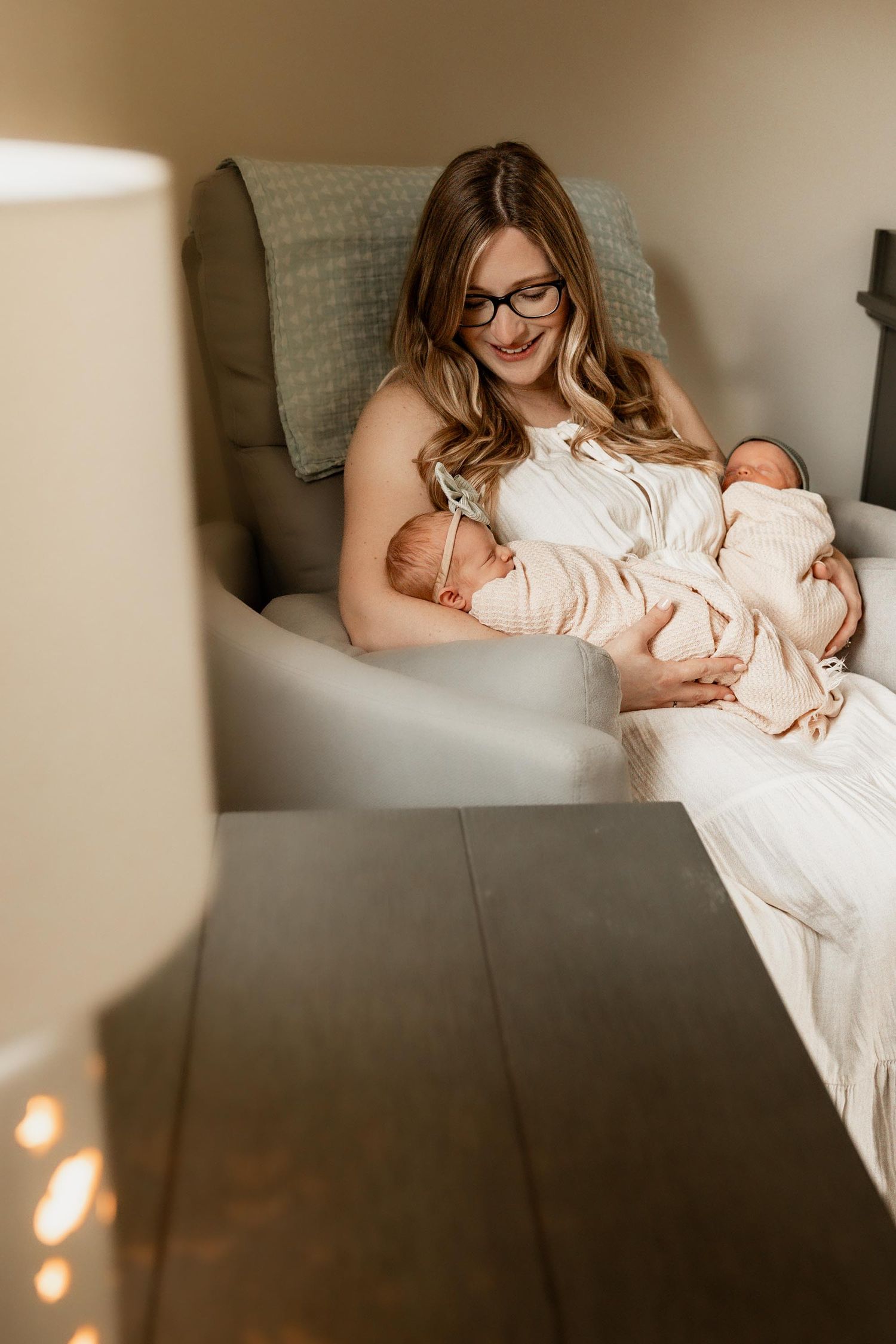 A smiling mother with glasses sits in a gray chair, lovingly holding her twin newborn babies wrapped in soft pink blankets.