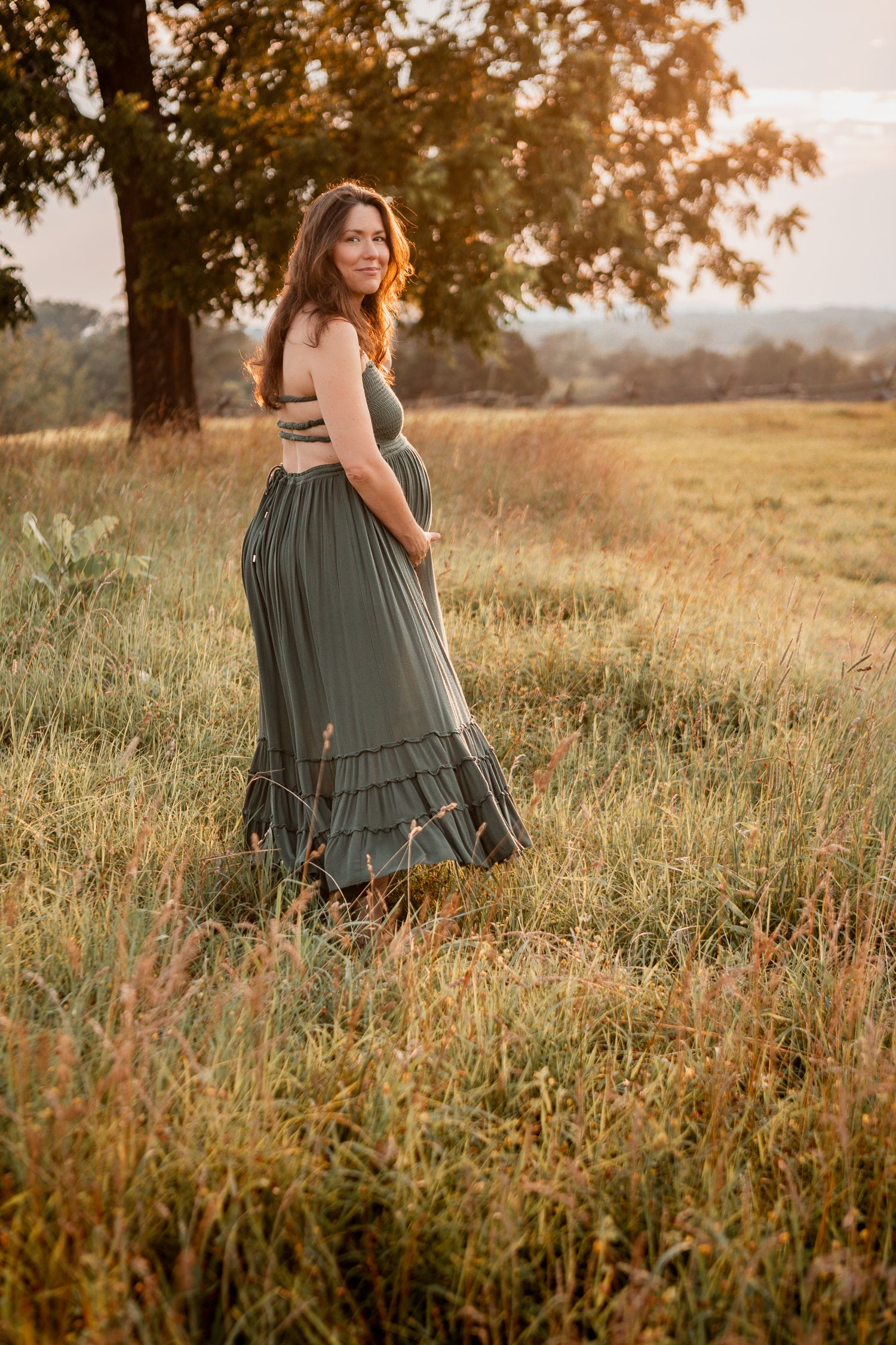 A person in a flowing sage green dress stands in a golden field at sunset while touching their stomach.