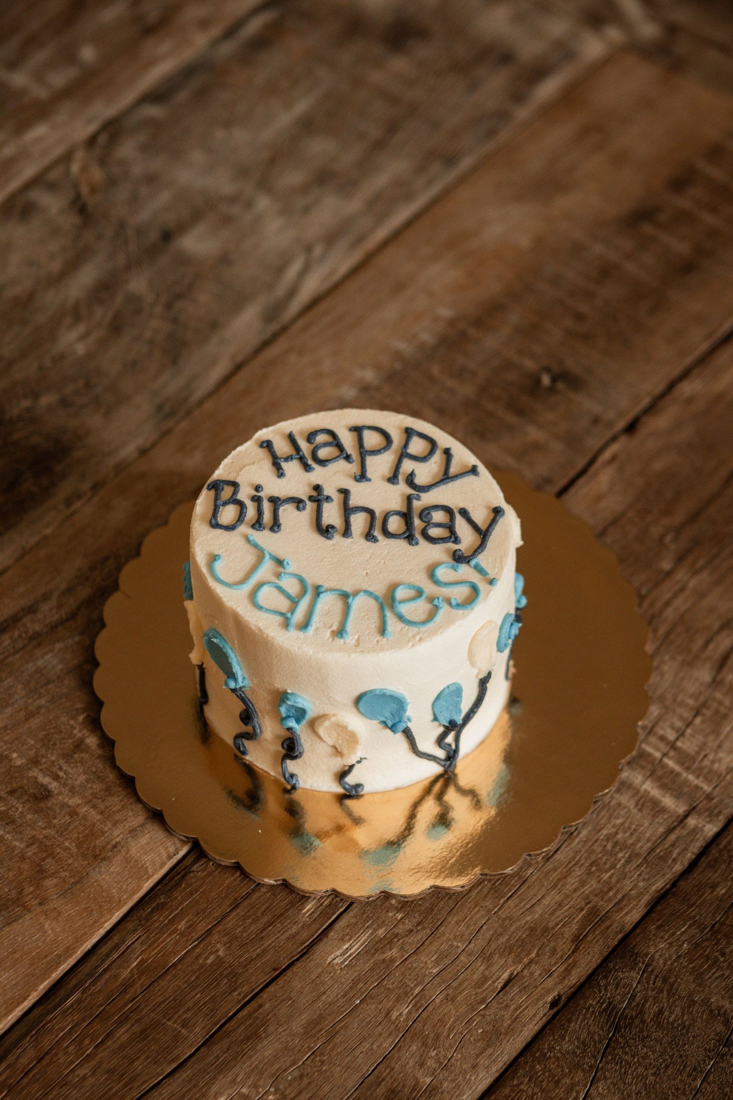 A small white birthday cake with blue decorative details and 'Happy Birthday' text sits on a rustic wooden surface.