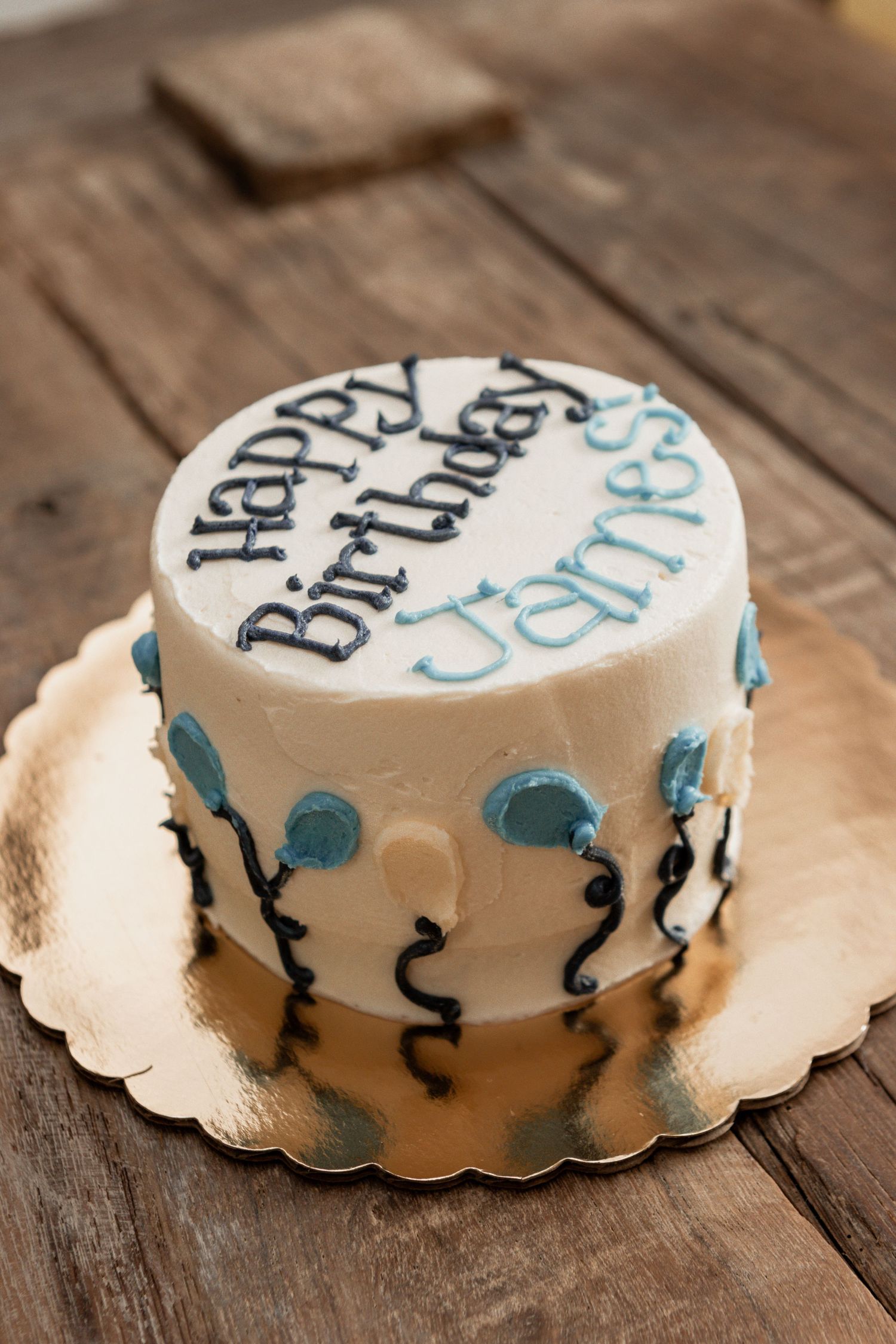 White birthday cake with black and turquoise lettering seen from multiple angles on rustic wooden table.