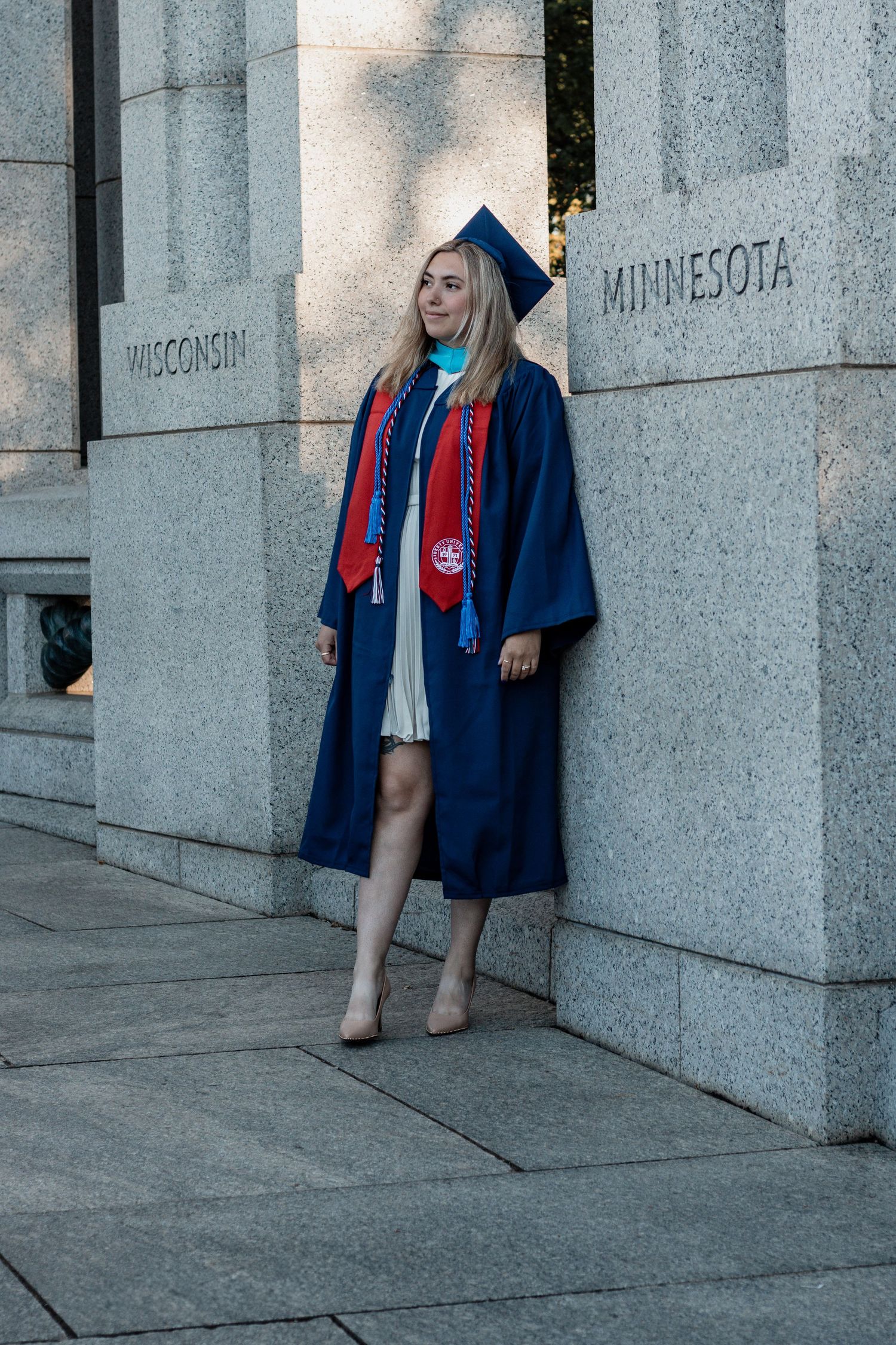 Graduate in blue cap and gown poses against stone pillars marked Wisconsin and Minnesota during commencement.