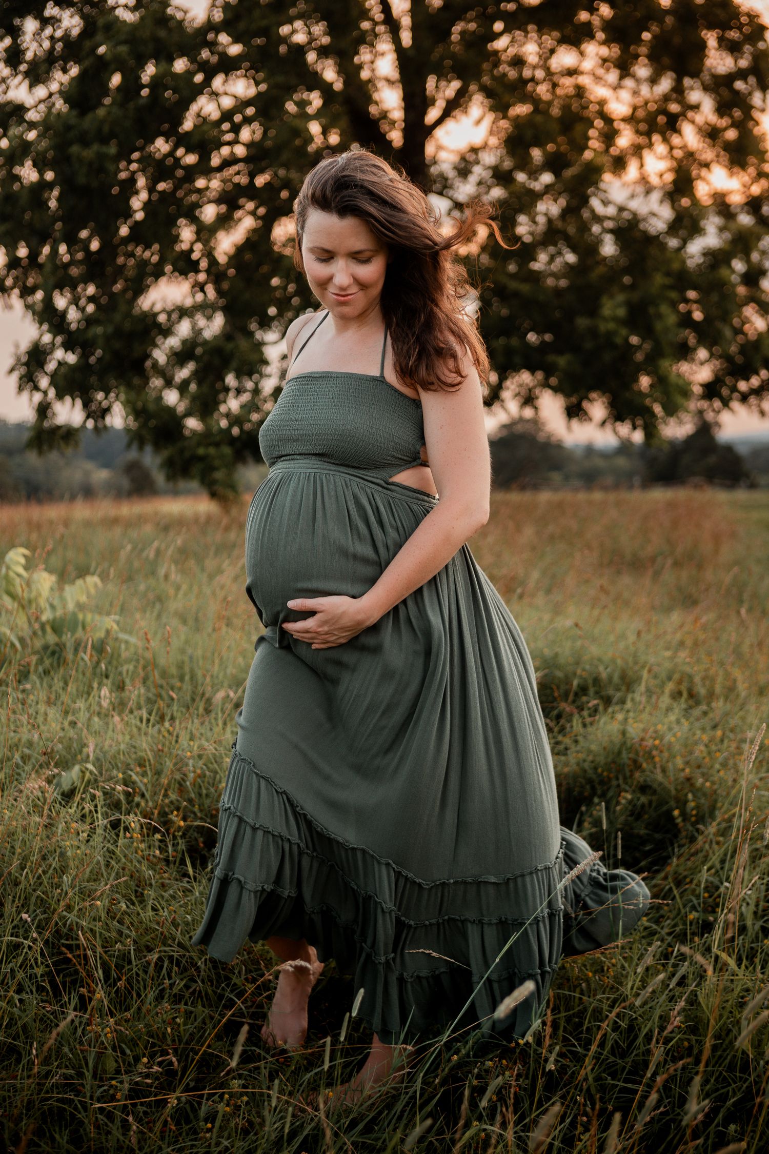 Pregnant woman in flowing green dress stands in golden field at sunset with wind blowing through her hair.