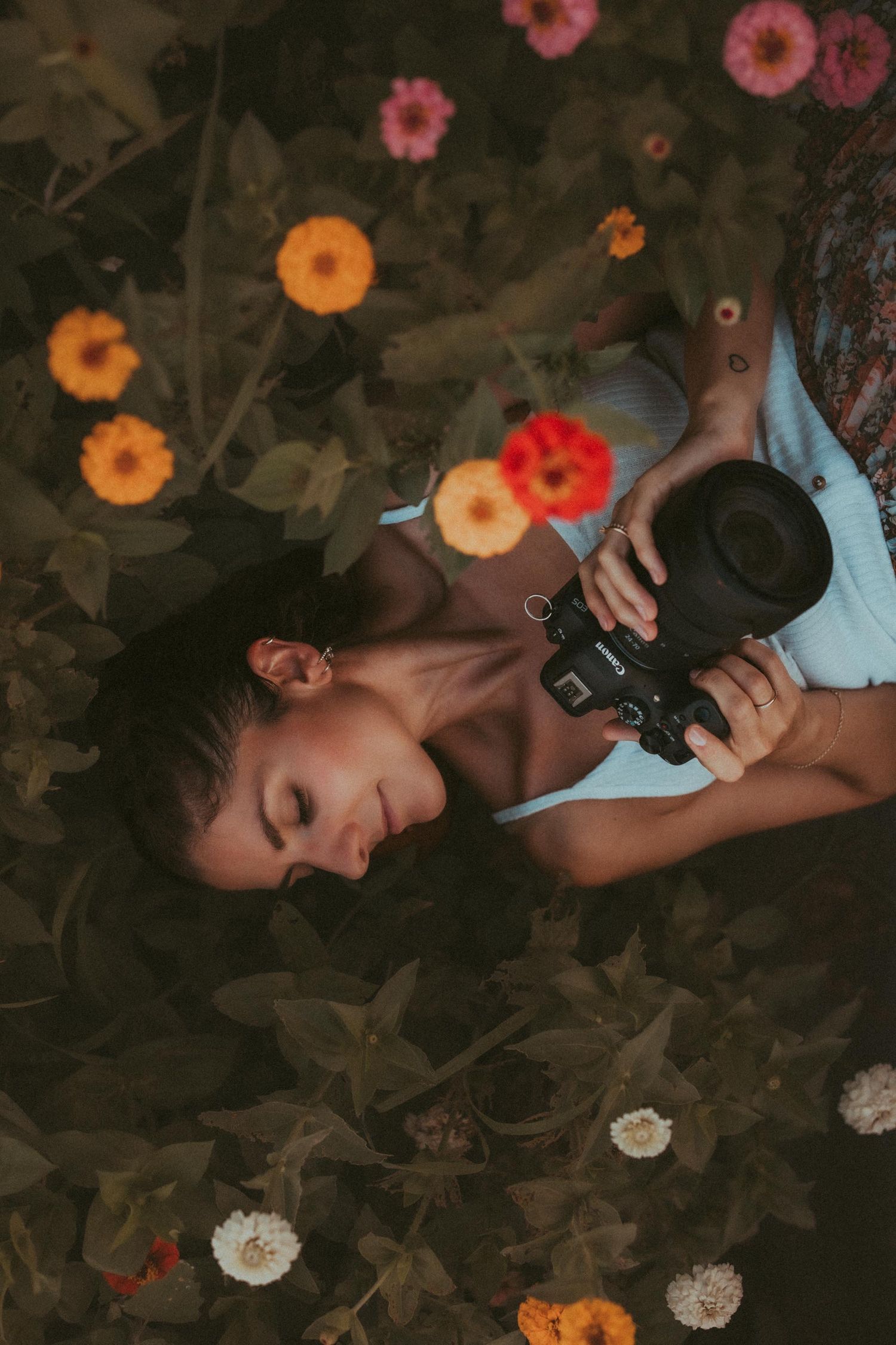 Young woman lying among colorful zinnia flowers in a garden, holding a vintage camera with eyes closed.