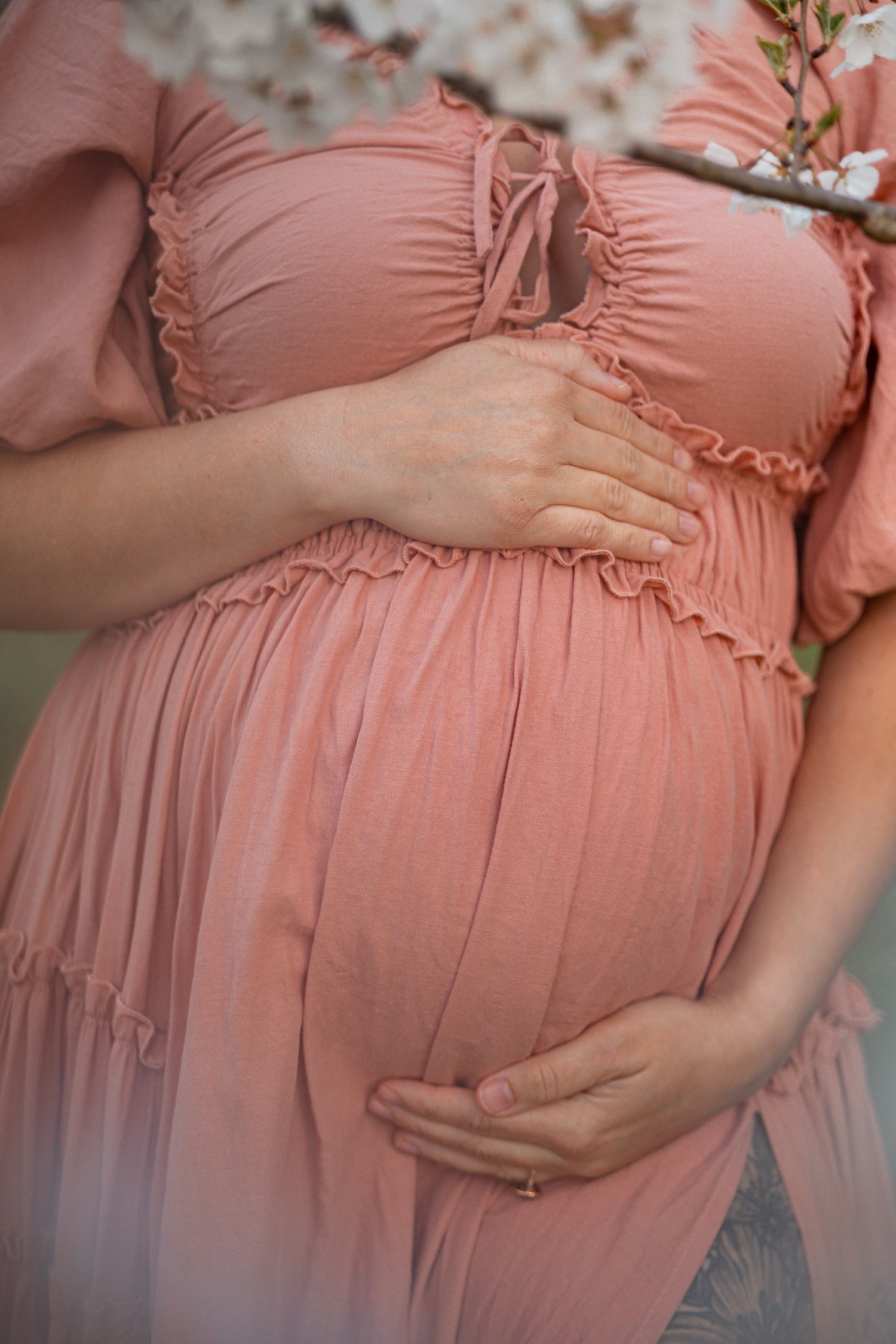 Pregnant woman in pink ruched dress cradling baby bump, maternity photo with cherry blossoms.