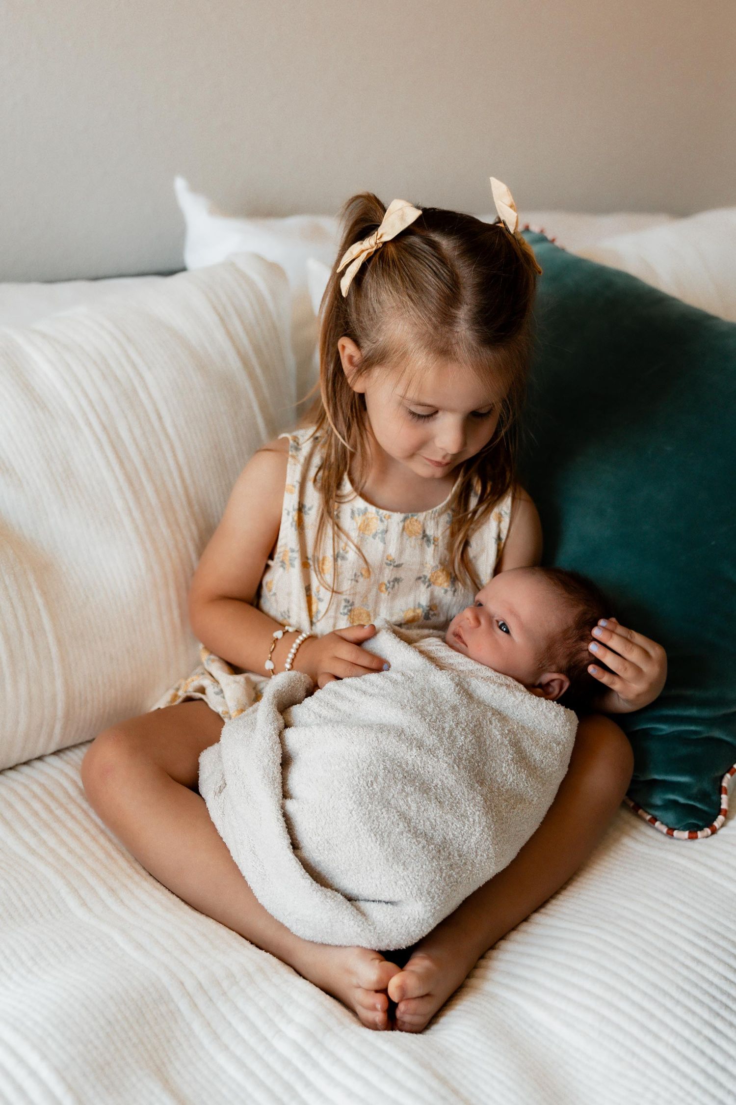 Young girl with bow sits on bed lovingly holding swaddled newborn baby sibling in soft white blanket.