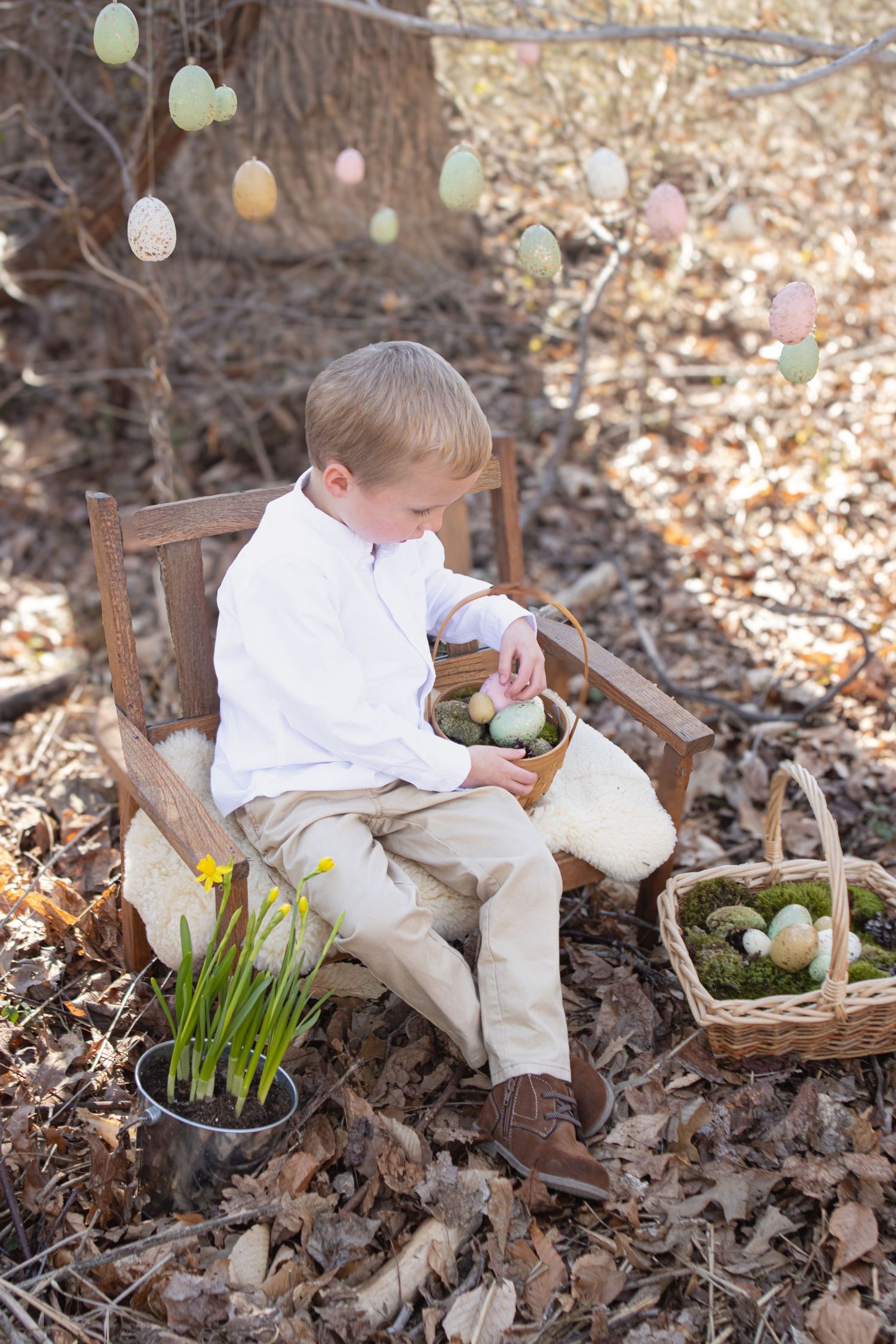Child in white shirt sits on wooden chair examining Easter basket with daffodils nearby.