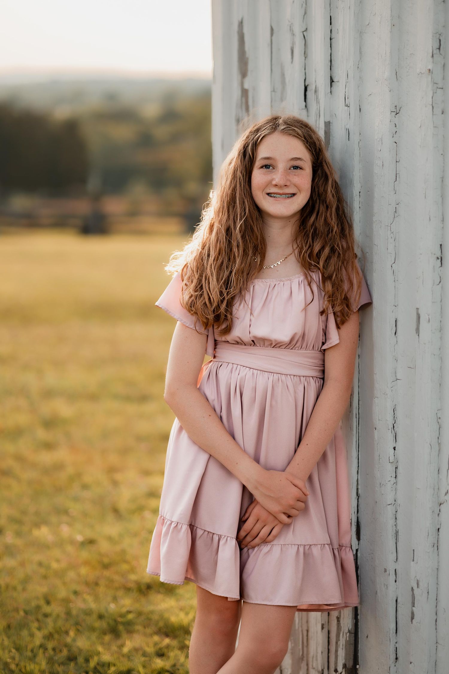 A teenage girl with curly hair in a pink dress leans against a white barn at golden hour.