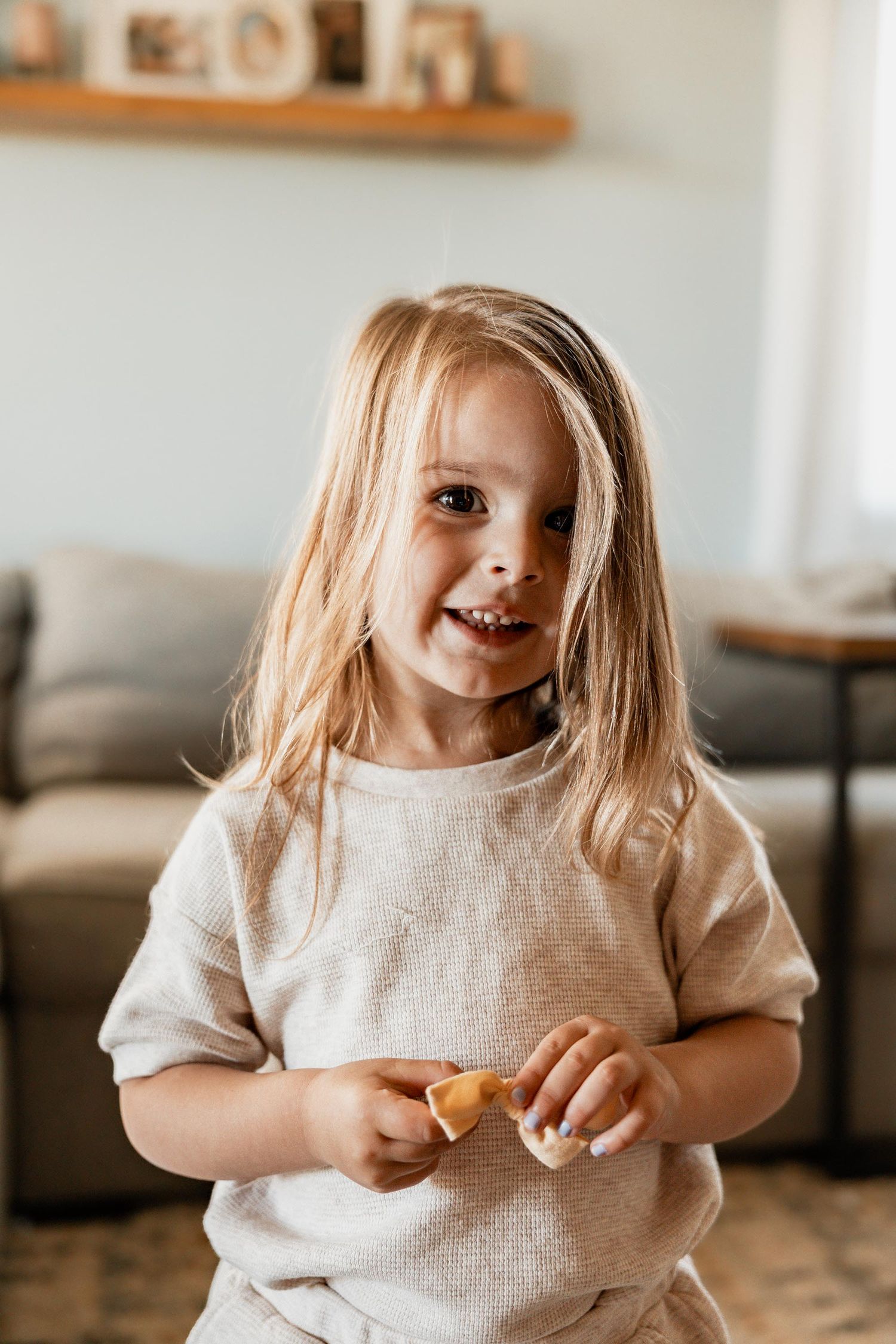 Smiling young girl with blonde hair holding a cookie in a cozy living room setting.