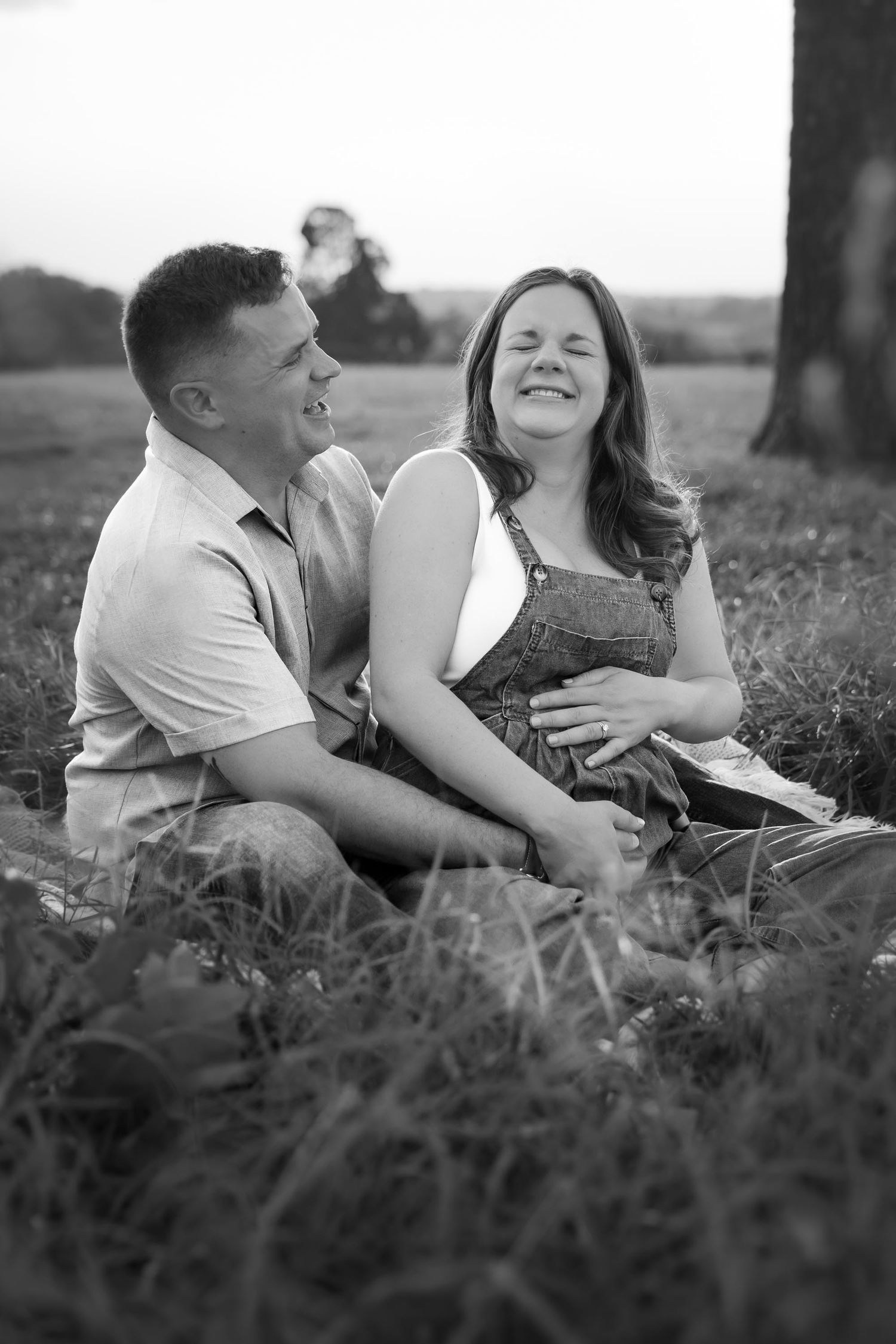 Black and white photo of a laughing couple sitting together in a grassy field, sharing a joyful moment outdoors.