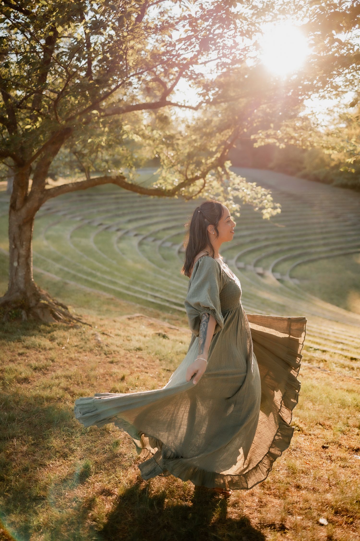Woman in flowing sage green dress twirling in golden sunlight near a tree with terraced green hills backdrop.