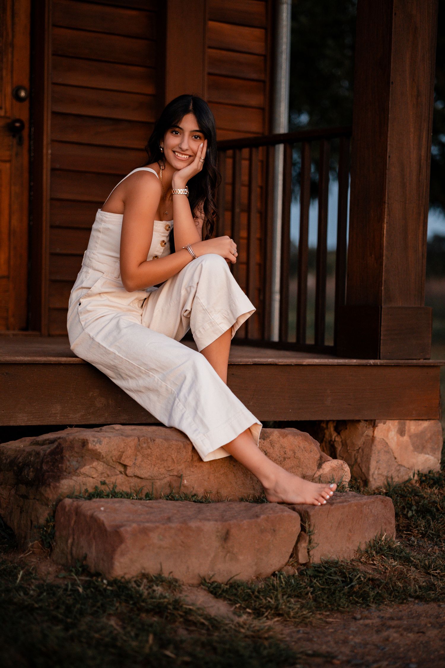 Person in white dress sitting on rustic stone steps by wooden railing at dusk.