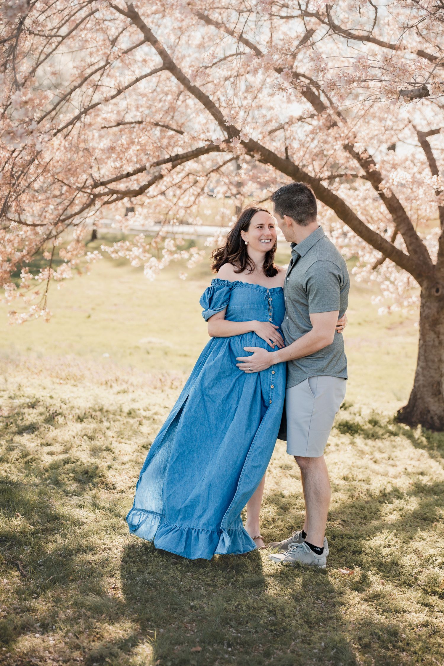 Pregnant couple embracing under blooming cherry blossom trees, woman in blue off-shoulder dress, man in gray shirt.