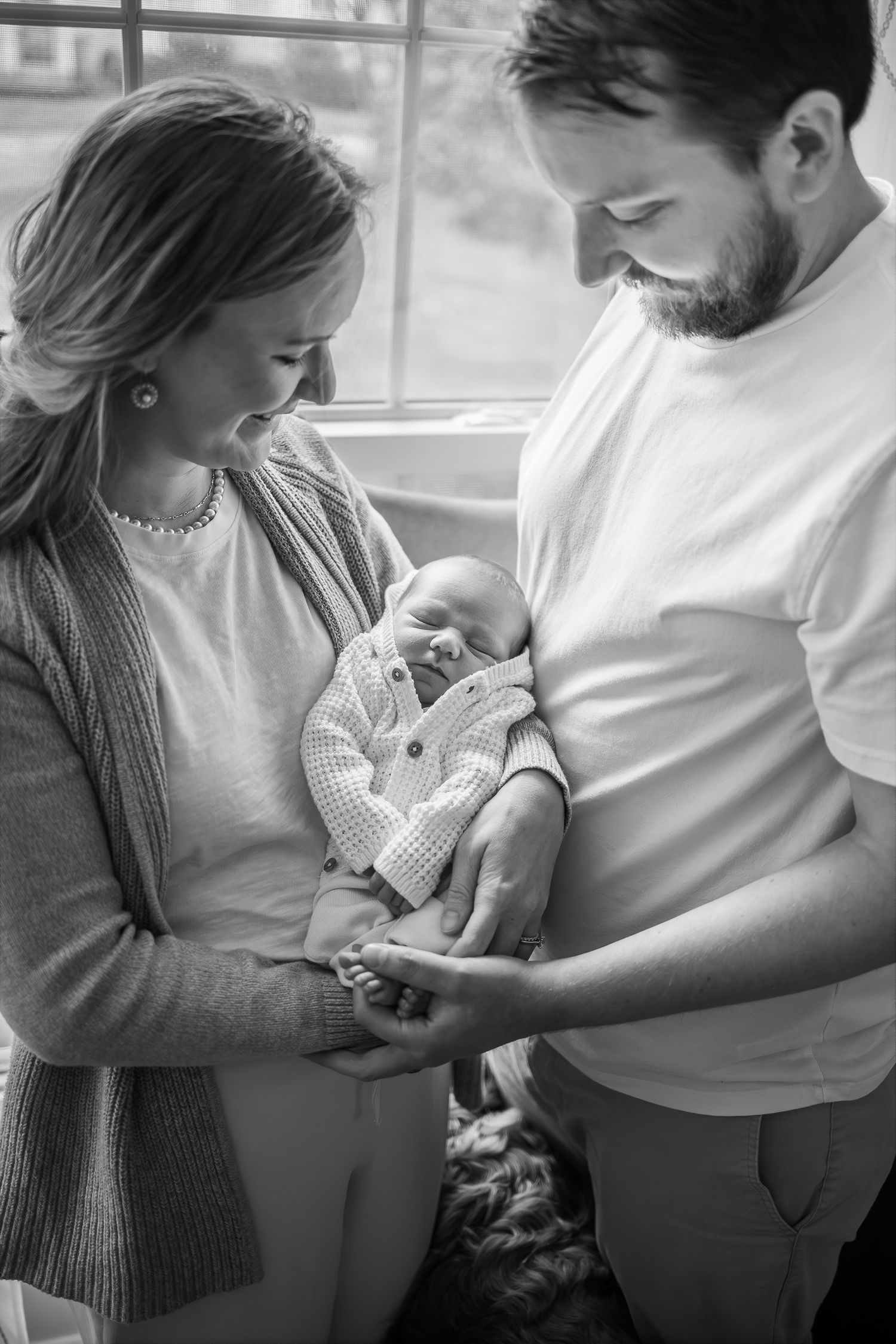 Black and white photo of couple holding newborn baby by window.