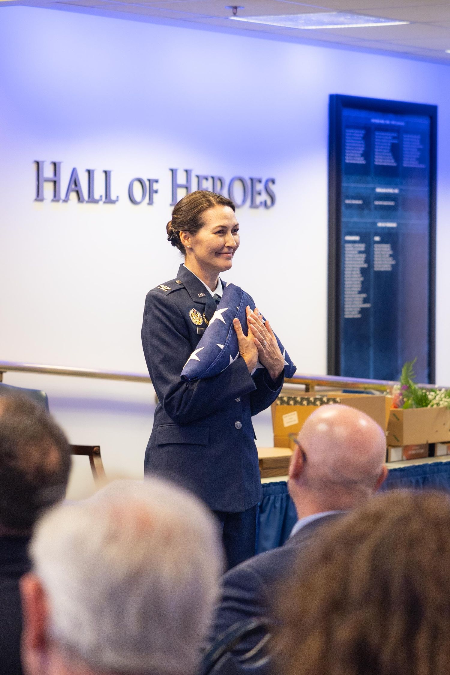 Female military officer in uniform holds folded flag while speaking at Hall of Heroes ceremony.