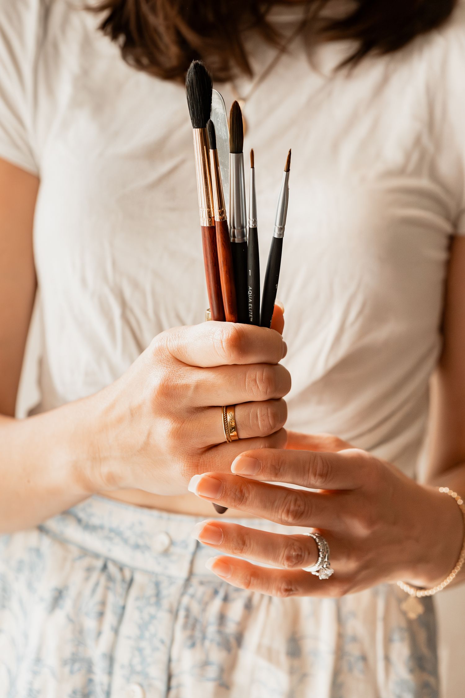 Hands wearing rings hold a collection of artist paint brushes against a white shirt and floral skirt.