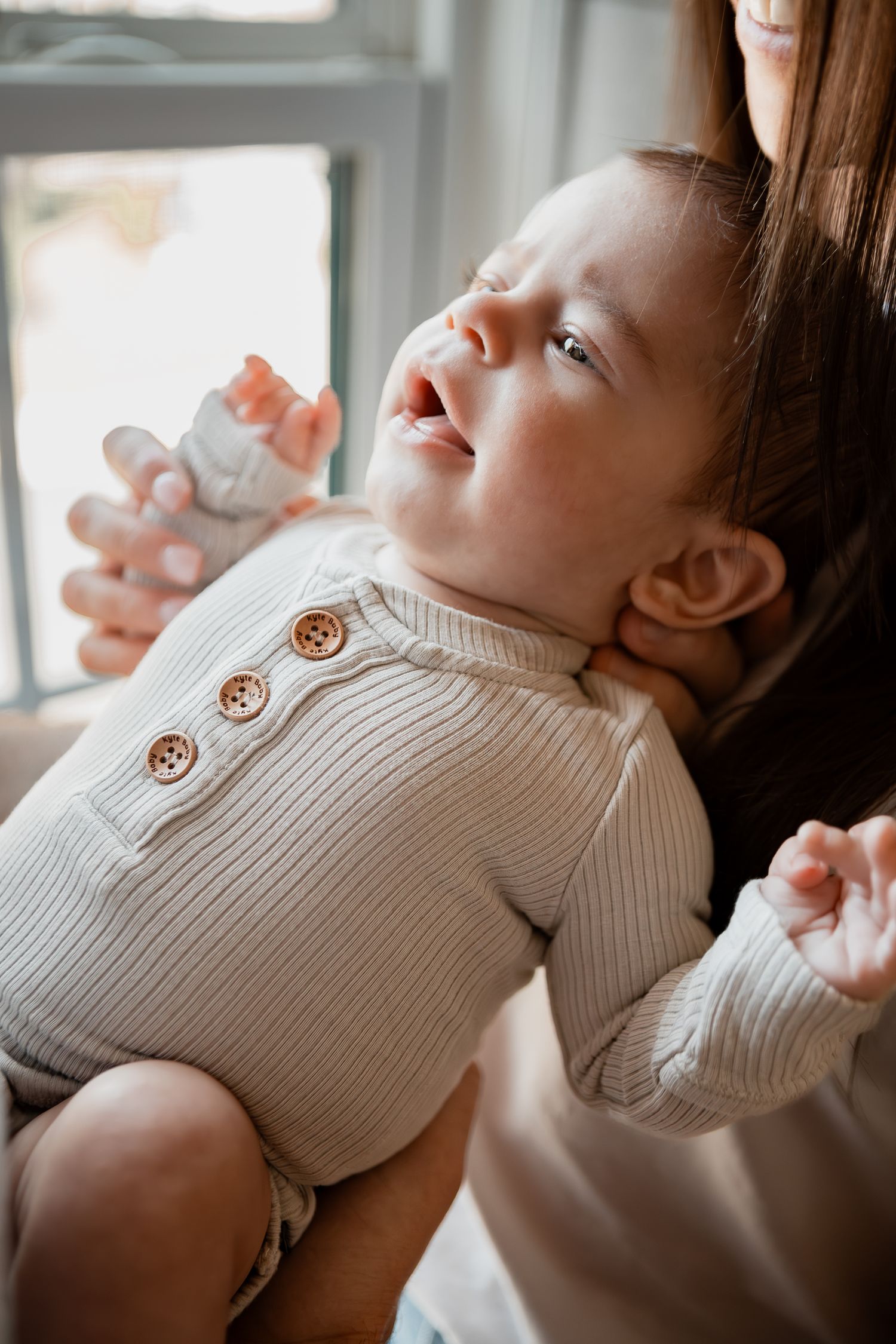 A close-up series showing a baby in a white ribbed romper with wooden buttons laying back against a window.