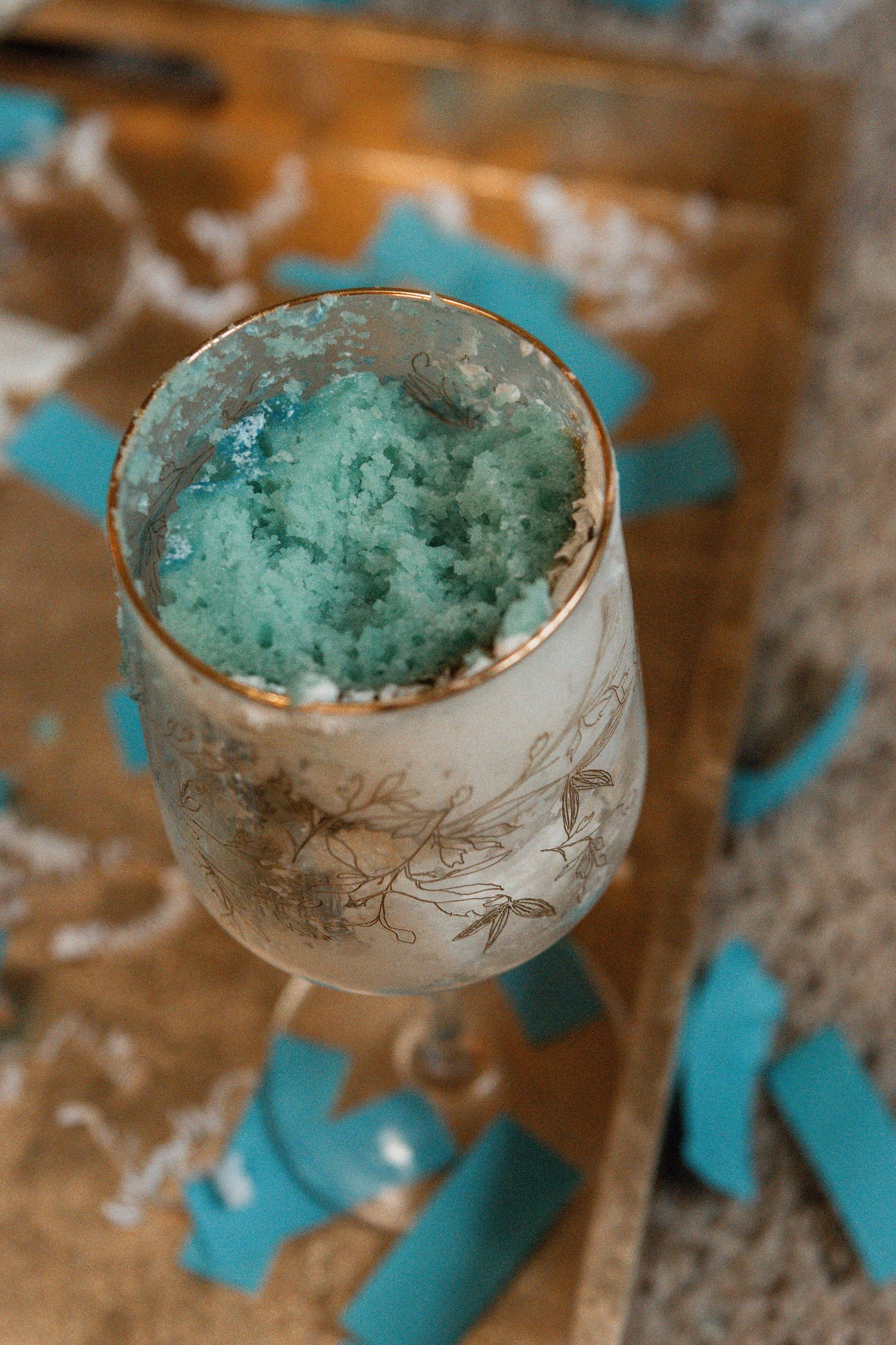A frosted white wine glass with a blue-green icy slushy mixture inside sits on a rustic wooden surface with teal accents.