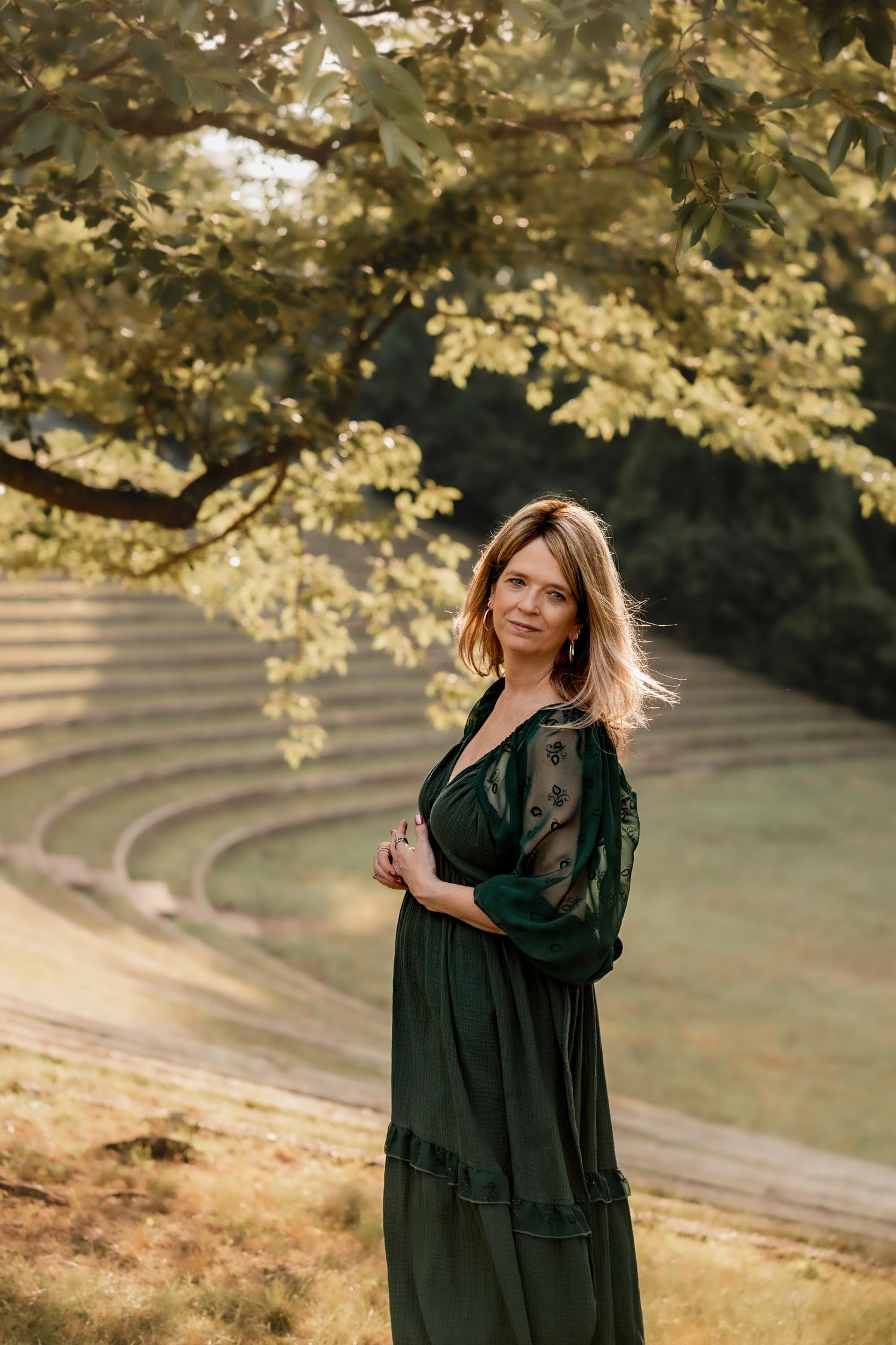 Woman in elegant green dress standing under a tree in a park with stone amphitheater steps in background.